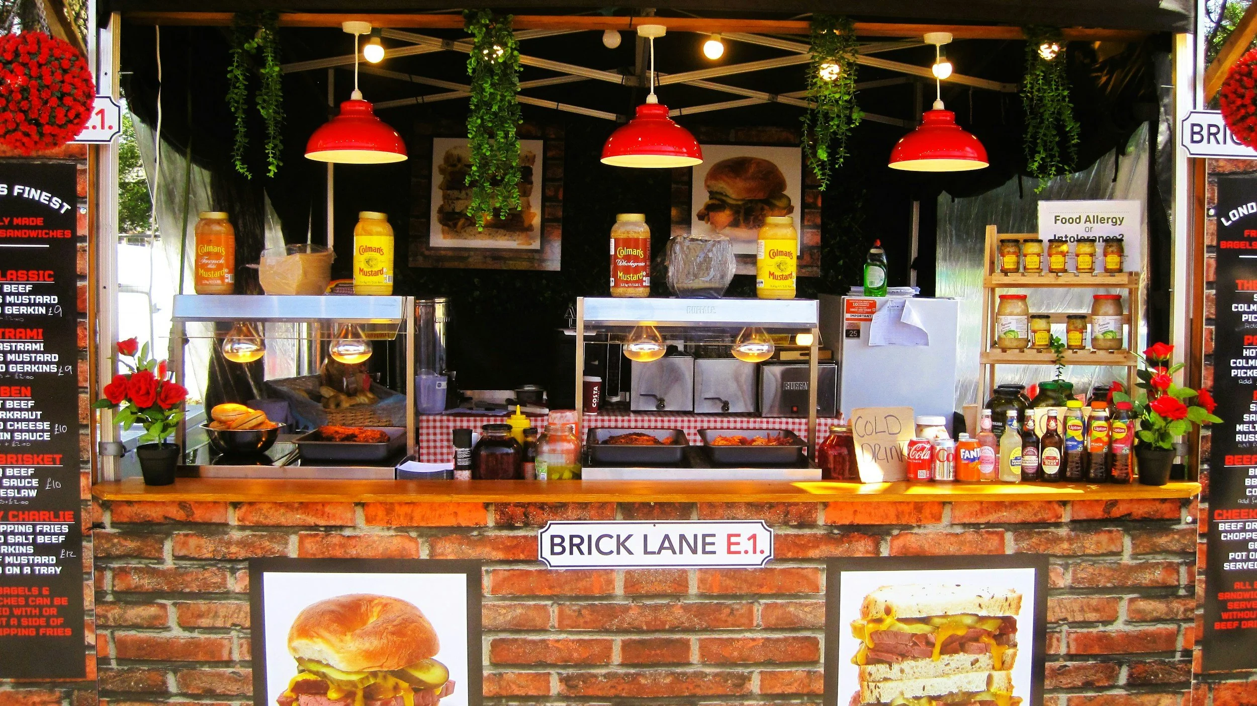 Carrito de comida de estilo feria con ladrillos y decoraciones verdes, con anuncios de comida en los lados, varias botellas y frascos en la barra, y cartel que dice 'BRICK LANE E.1'.