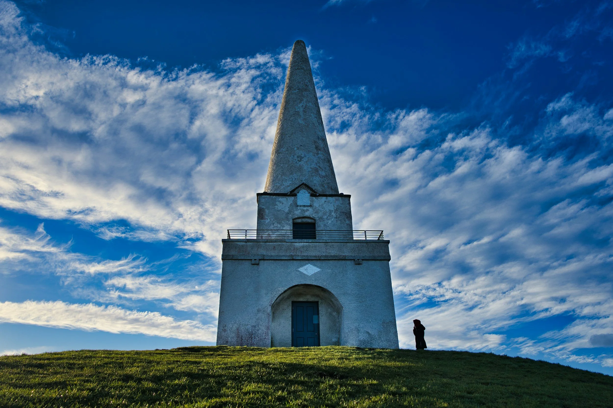 Torre blanca en una colina con cielo azul y nubes, con una persona de pie a la derecha.