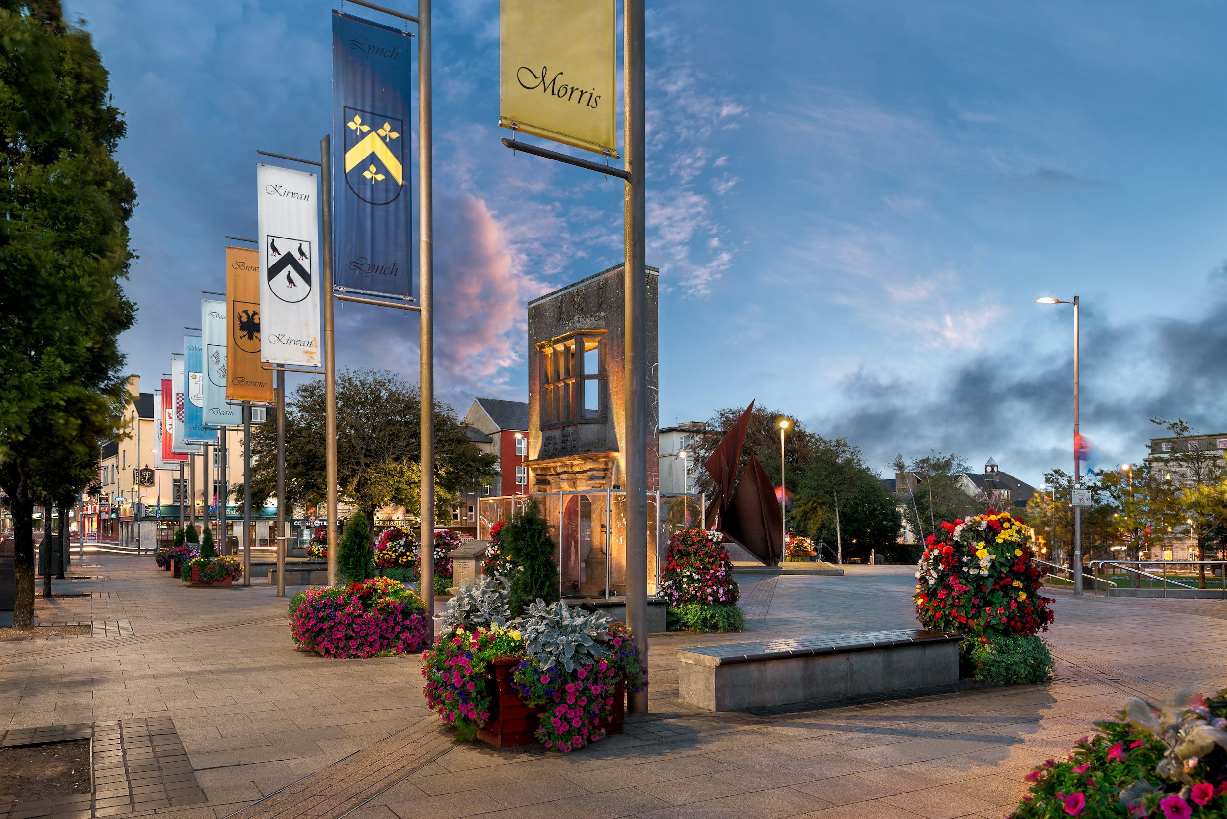 Plaza peatonal con banderas y flores de colores, edificio en el fondo y cielo parcialmente nublado al atardecer.
