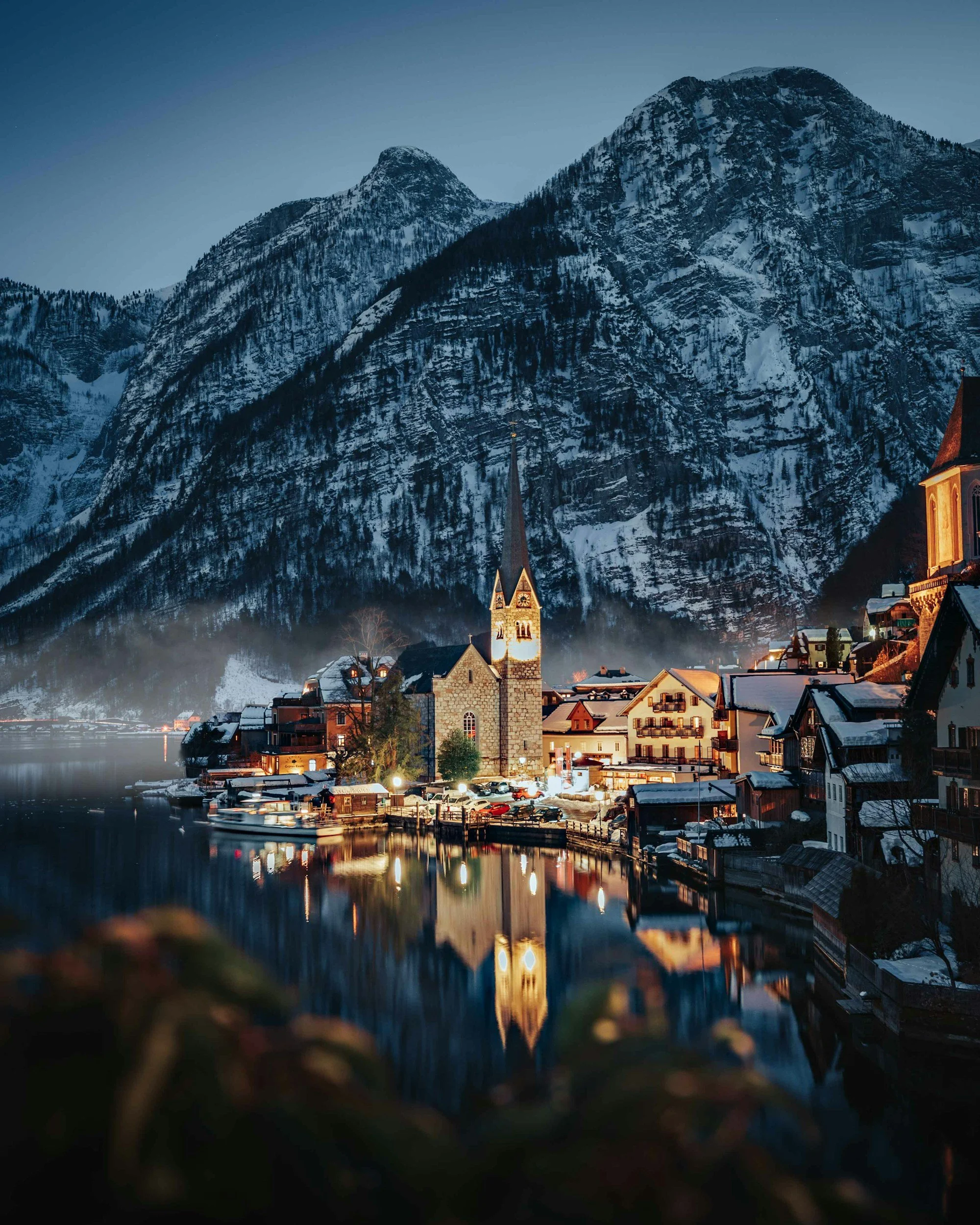 Pueblo junto a un lago con montañas nevadas al fondo, iluminado por luces nocturnas, con un centro de iglesia y reflejos en el agua.