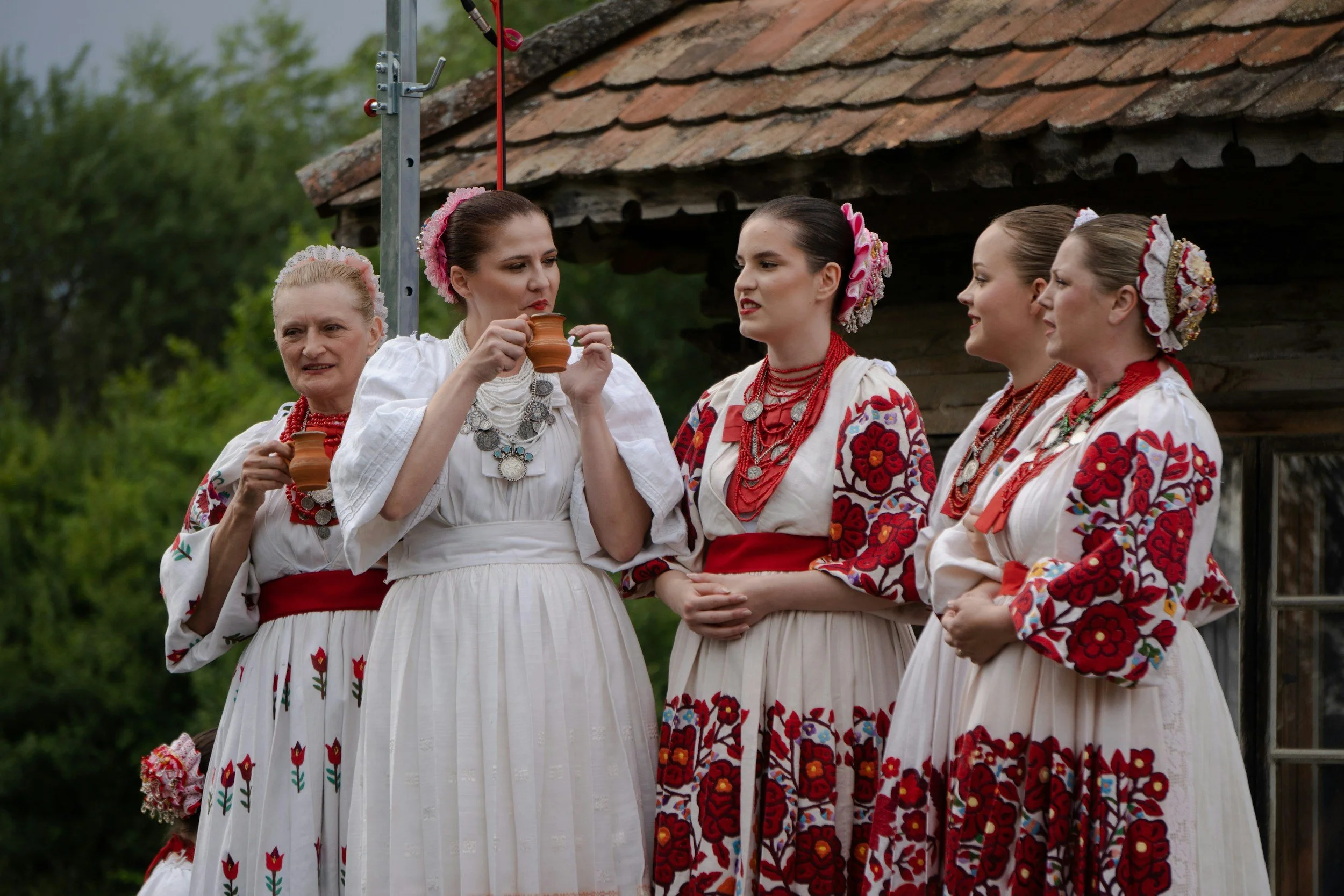 Cinco mujeres vestidas con trajes tradicionales folclóricos, con detalles de bordados y accesorios de joyería, participando en una celebración cultural al aire libre.