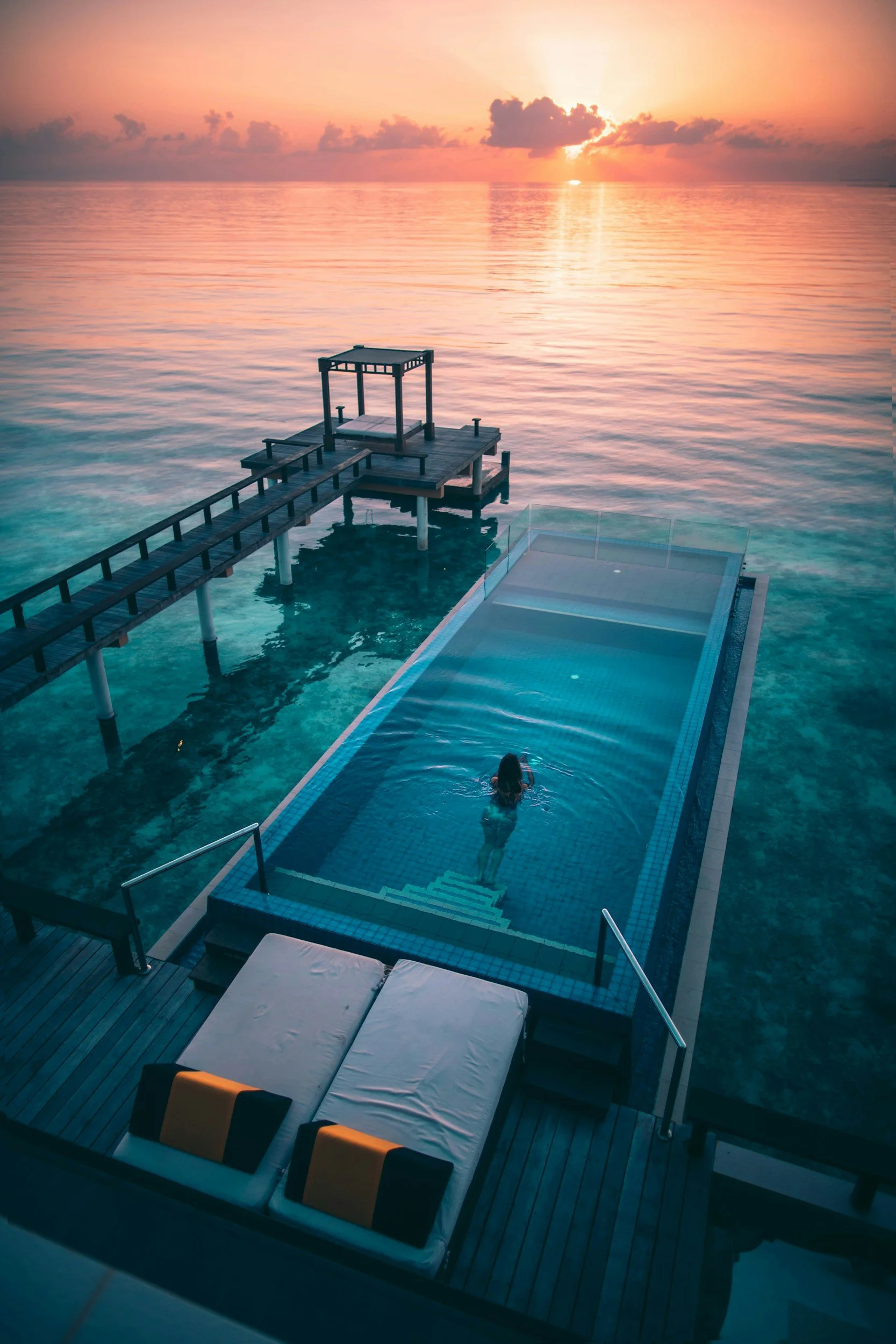 Piscina en un resort junto al mar durante una puesta de sol, con una persona nadando en la piscina y un muelle de madera que lleva a una estructura en el agua.