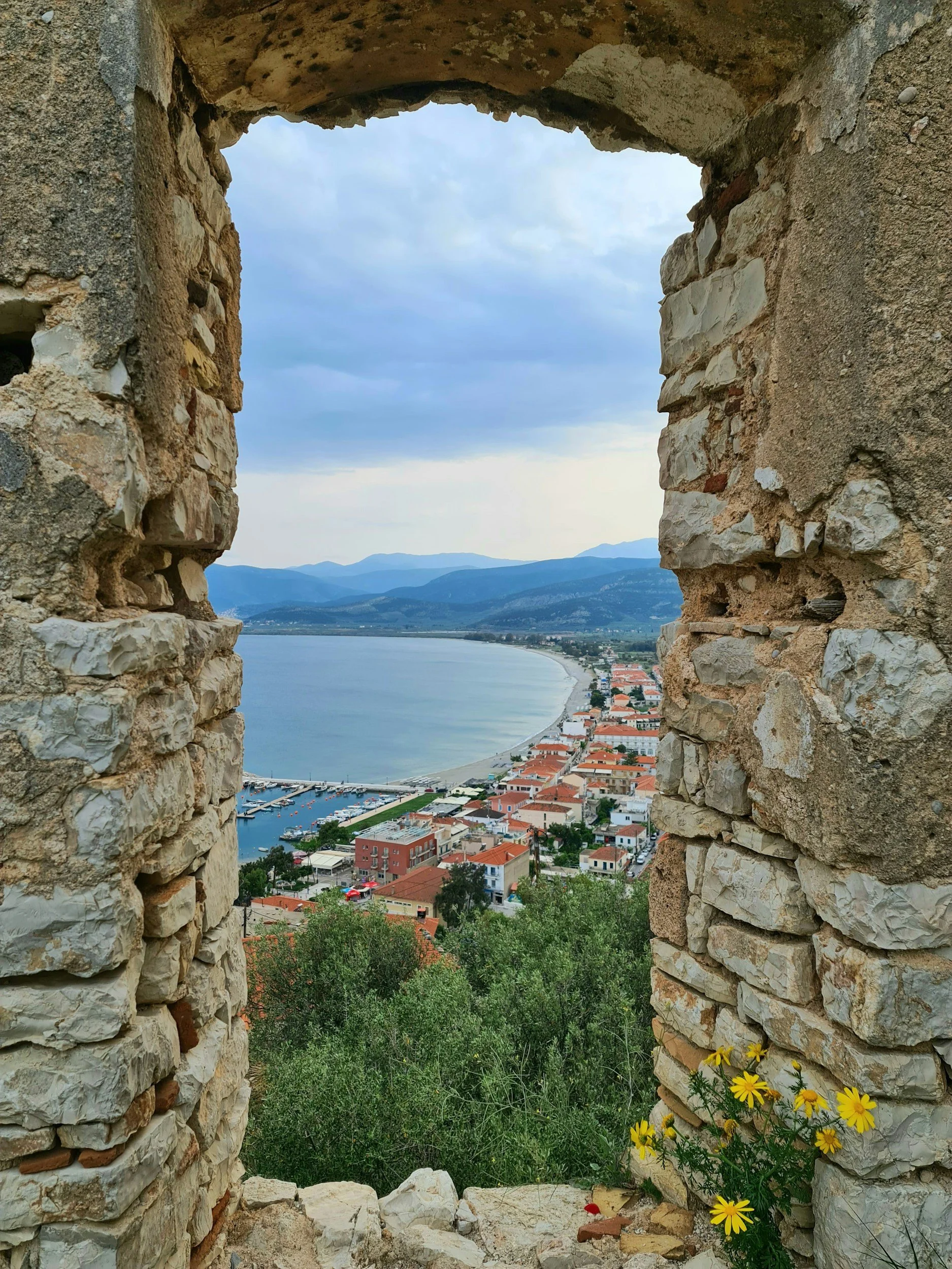 Vista de un pueblo costero a través de una ventana en ruinas, con mar, montañas y un cielo nublado.