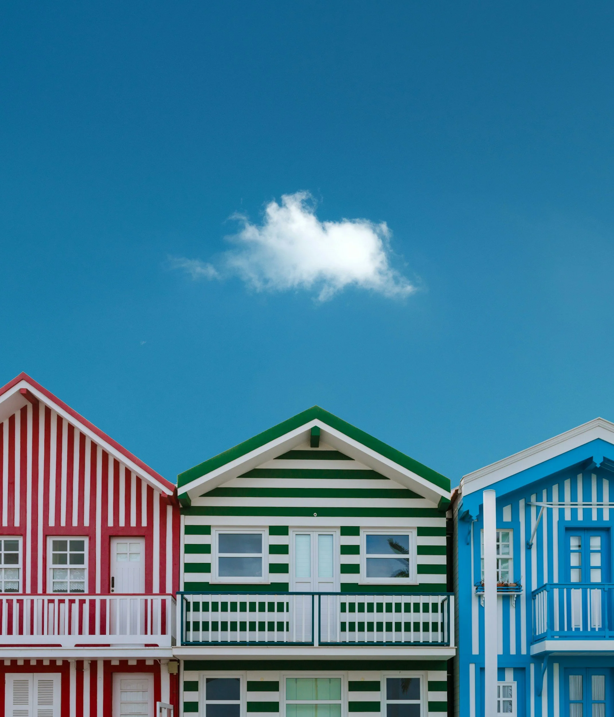 Tres casas coloridas con patrones de rayas en la fachada, una roja a la izquierda, una verde en el centro y una azul a la derecha, bajo un cielo azul con una nube blanca.