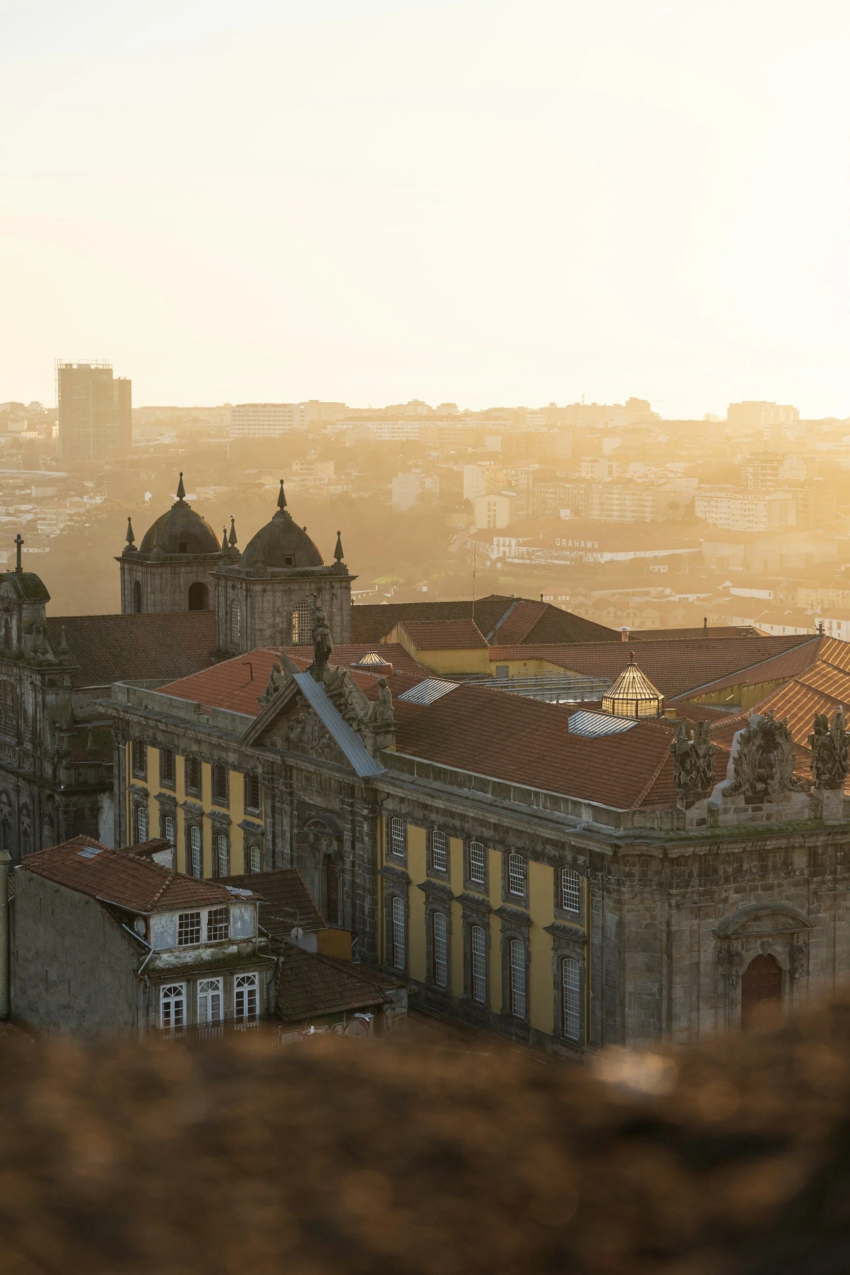 Vista de un edificio histórico con cúpulas y estatuas en la parte superior, con una ciudad moderna y el sol en el horizonte al fondo.