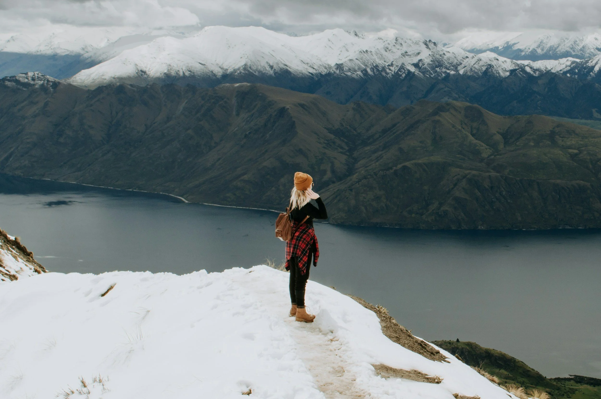 Mujer de espaldas en la nieve, observando un lago rodeado de montañas con picos nevados.