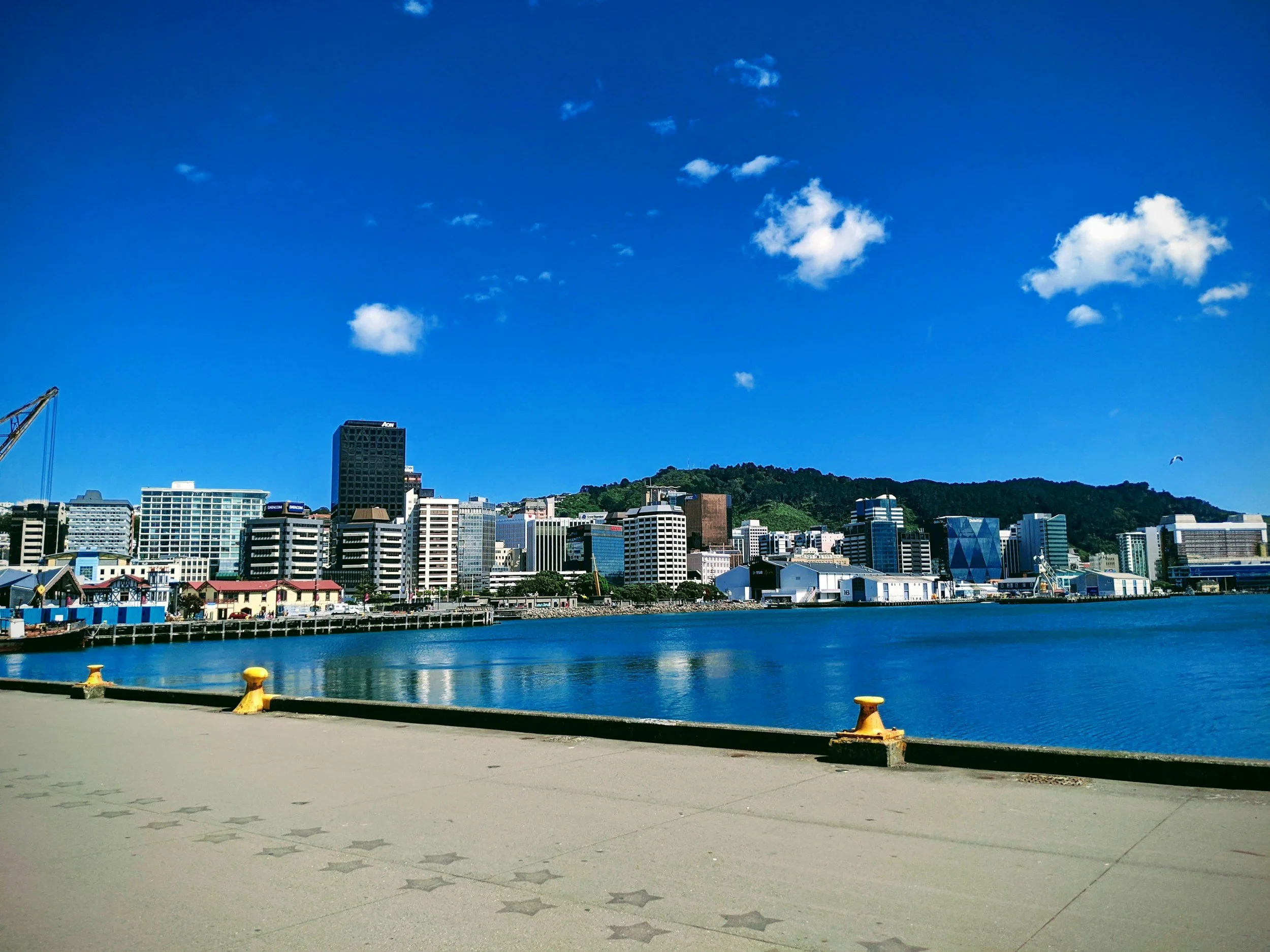 Vista de la ciudad con edificios modernos a la orilla del agua, con un cielo azul y algunas nubes, montañas en el fondo y un muelle en primer plano.