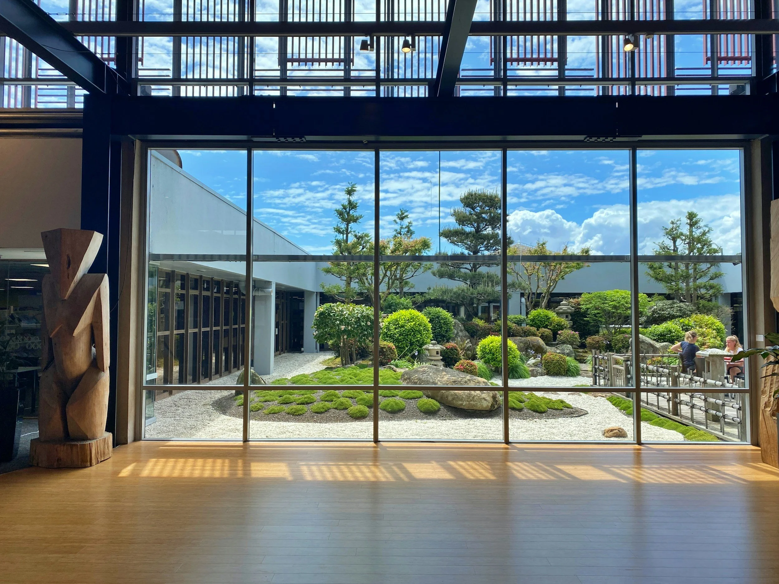Vista a través de un ventanal grande hacia un jardín japonés con plantas, rocas y caminos, en un día soleado con cielo azul y nublado.