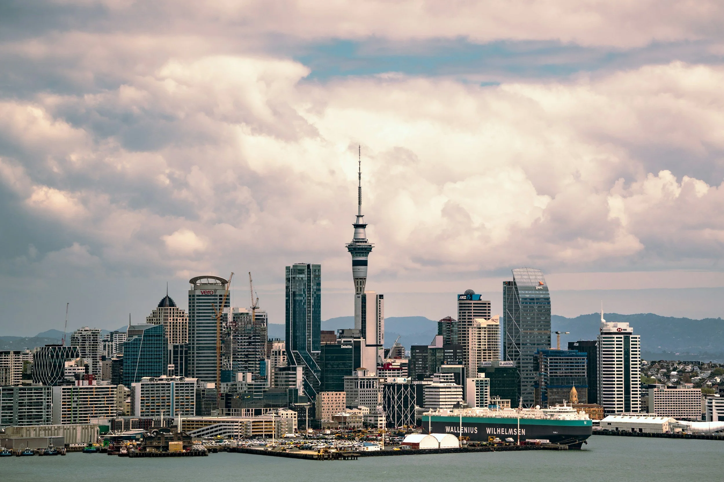Vista del skyline de Auckland, con la Torre de Auckland en el centro, edificios modernos y el puerto en primer plano bajo un cielo nublado.