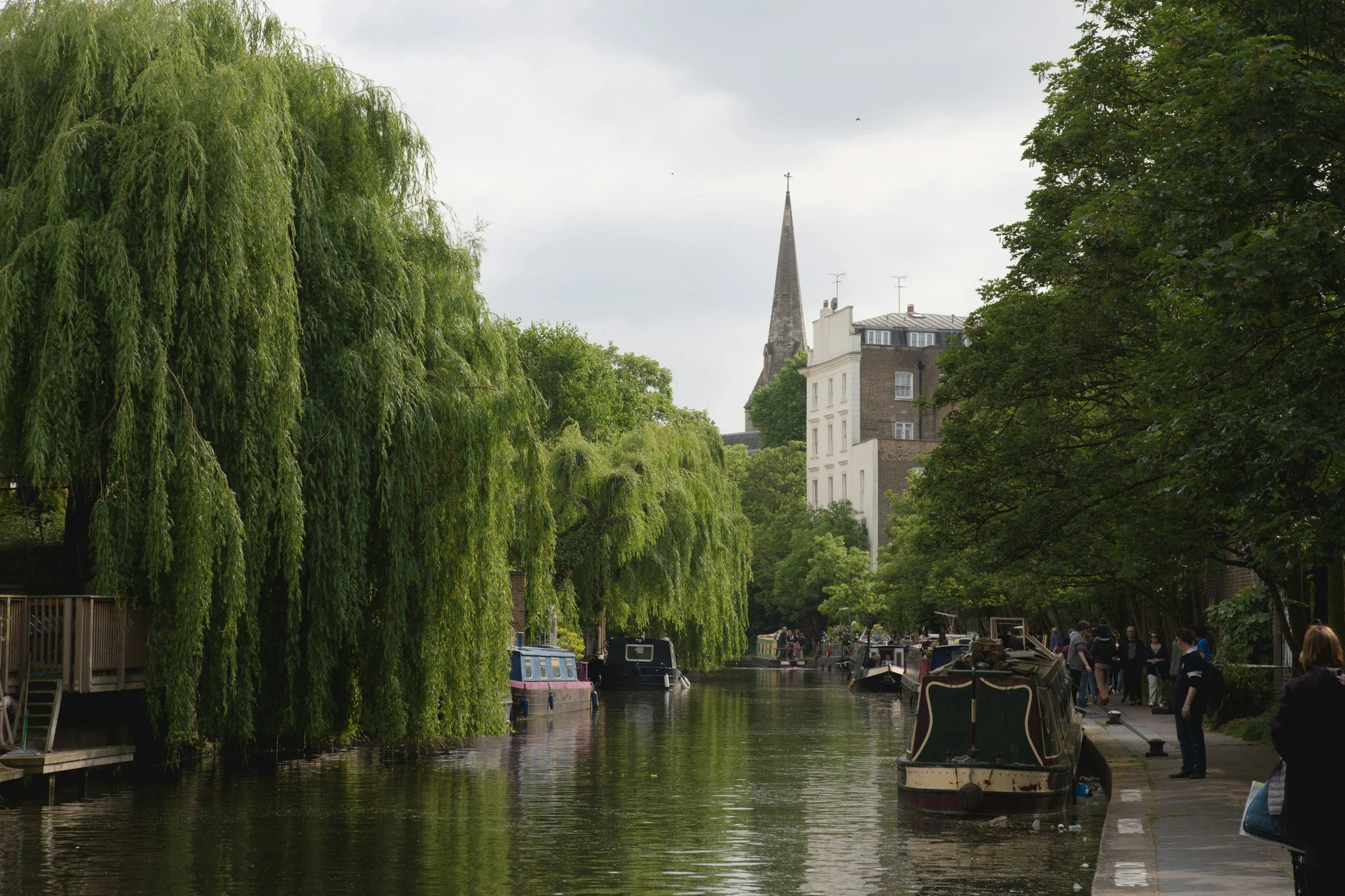 Canal con barcos y árboles verdes, edificios y una iglesia con torre en el fondo.