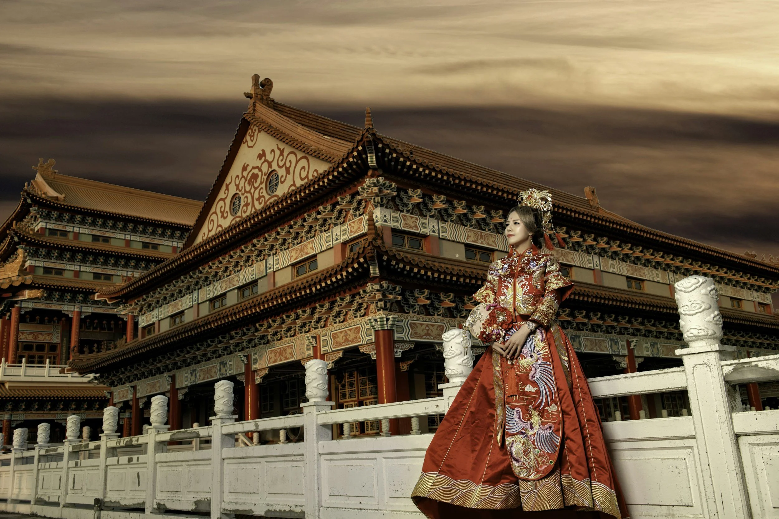 Una mujer vestida con ropa tradicional china, de color rojo y con detalles dorados, de pie frente a un edificio antiguo de estilo chino decorado con patrones y columnas rojas, con un cielo nublado al fondo.