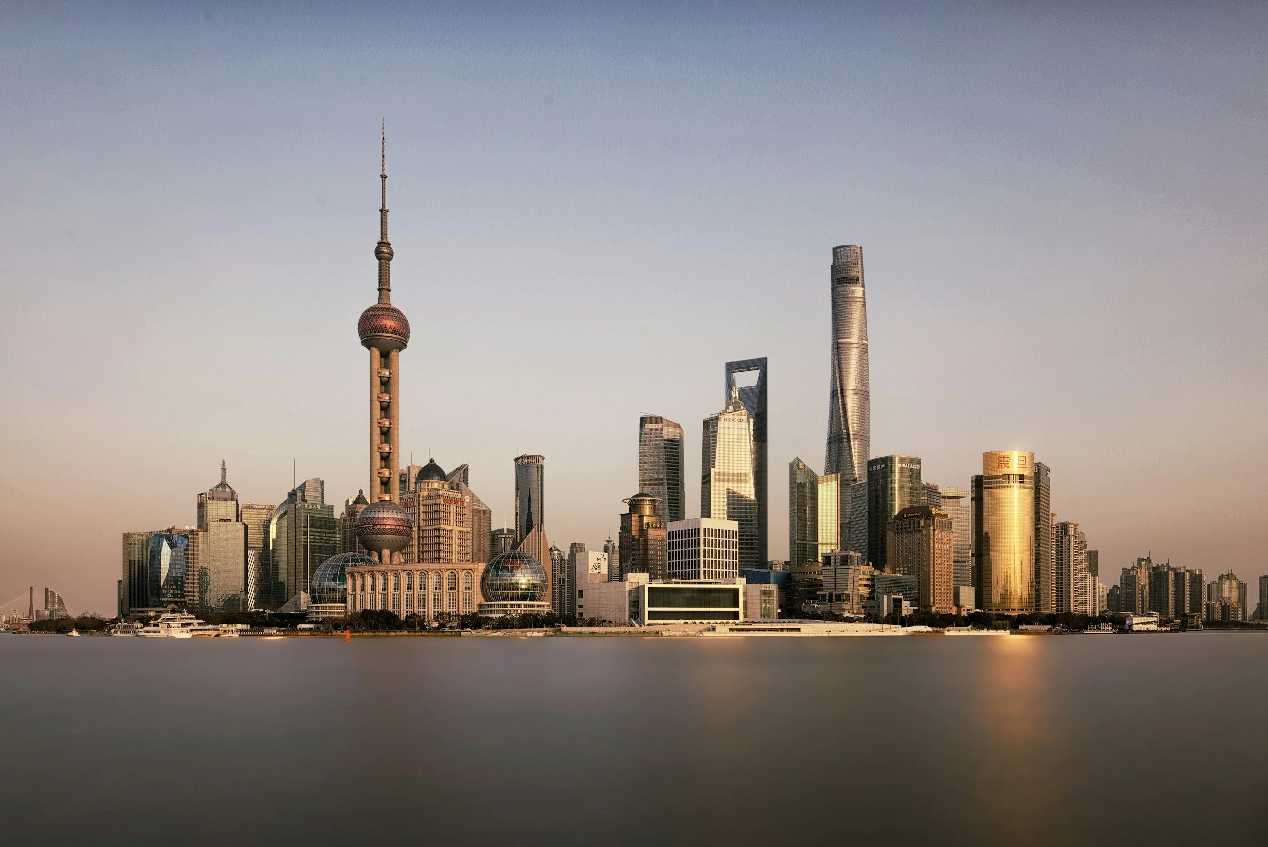 Vista del skyline de Shanghai con edificios como la Torre de Shanghai, la Torre de Shanghai y otros rascacielos modernos, reflejados en las aguas del río Huangpu en el atardecer.