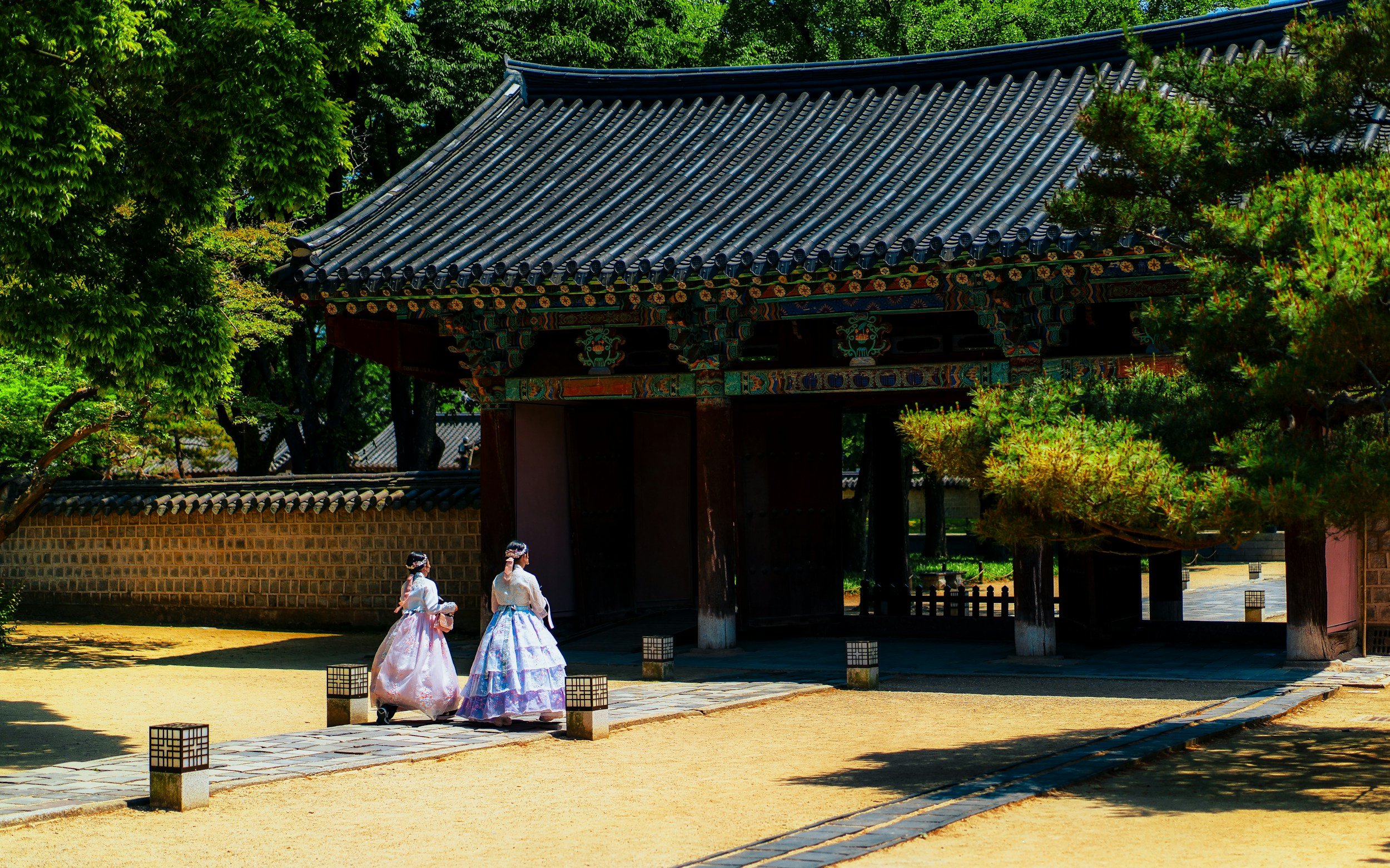 Dos mujeres vestidas con trajes tradicionales coreanos caminando cerca de una estructura antigua en un jardín con árboles verdes.