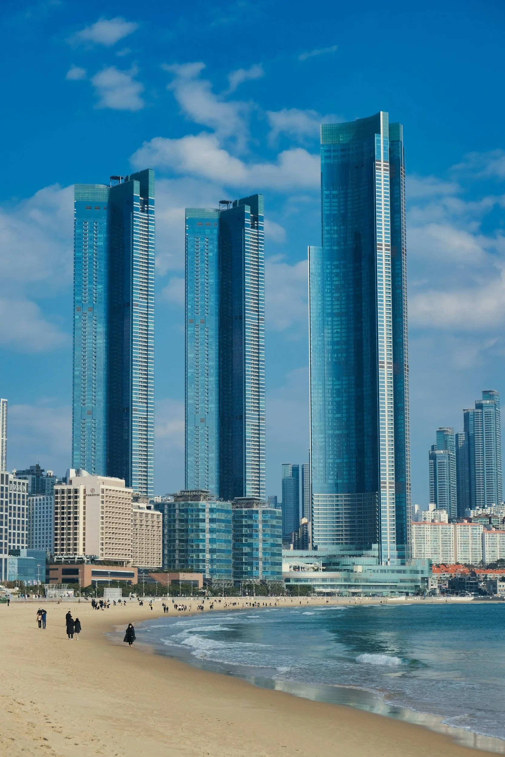 Edificios modernos de gran altura frente a la playa con arena y personas caminando, cielo azul con algunas nubes.