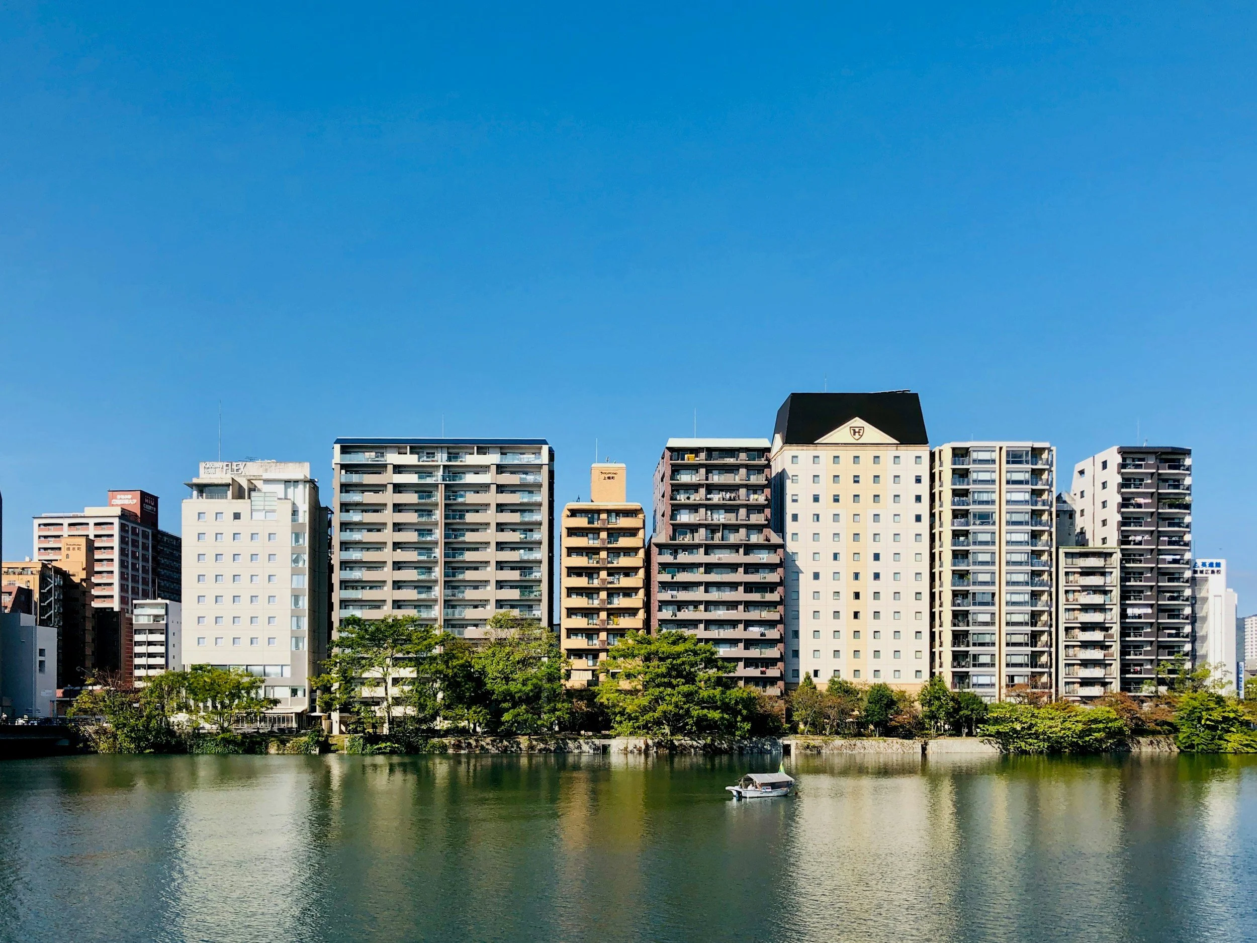 Paisaje urbano con edificios altos junto a un río, árboles y un cielo azul despejado