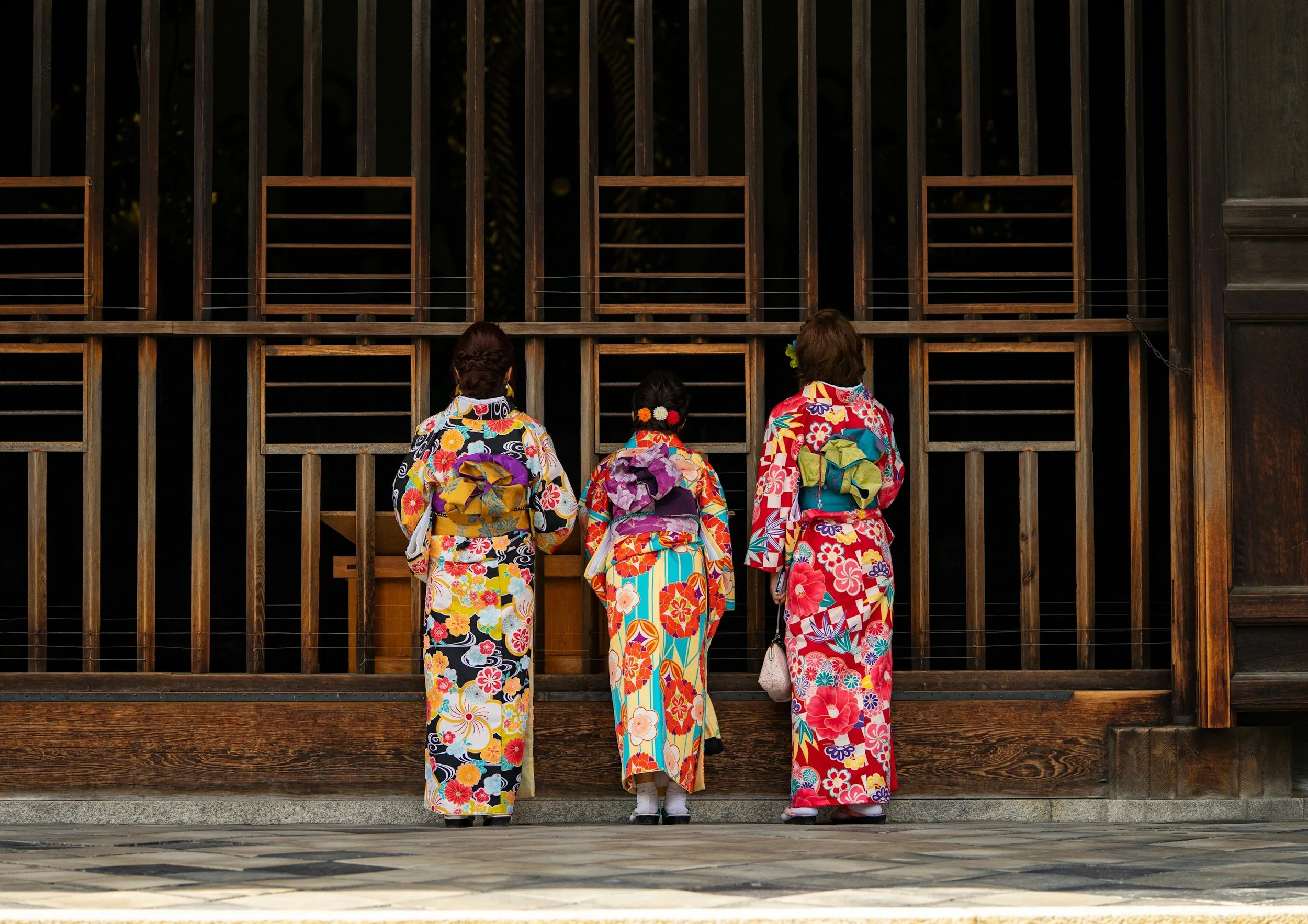 Tres mujeres vestidas con kimonos tradicionales japoneses observando a través de una estructura de madera en un templo o edificio antiguo.