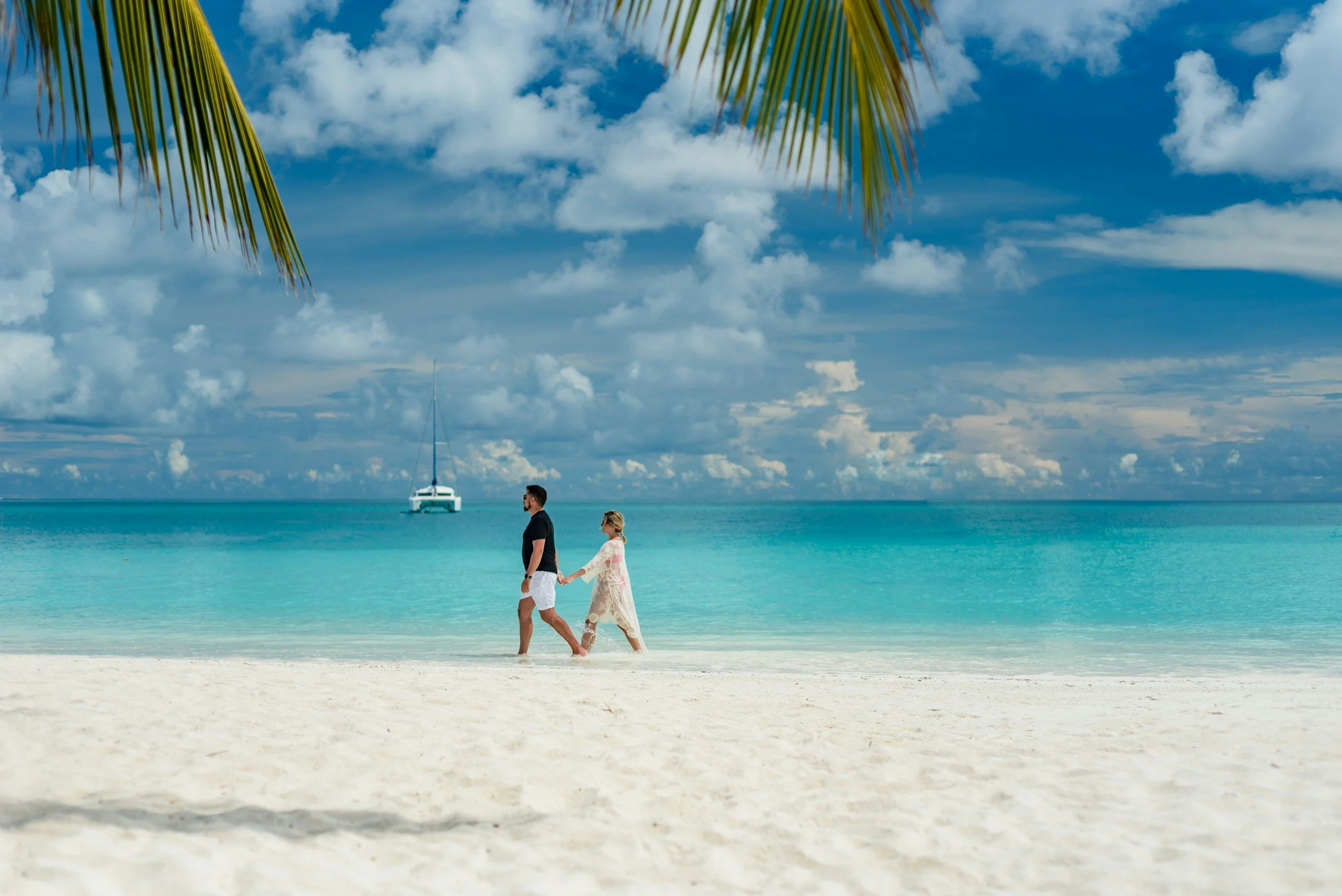 Una pareja caminando en la playa, con aguas turquesas y un barco en el fondo, bajo un cielo con nubes y palmera en primer plano.
