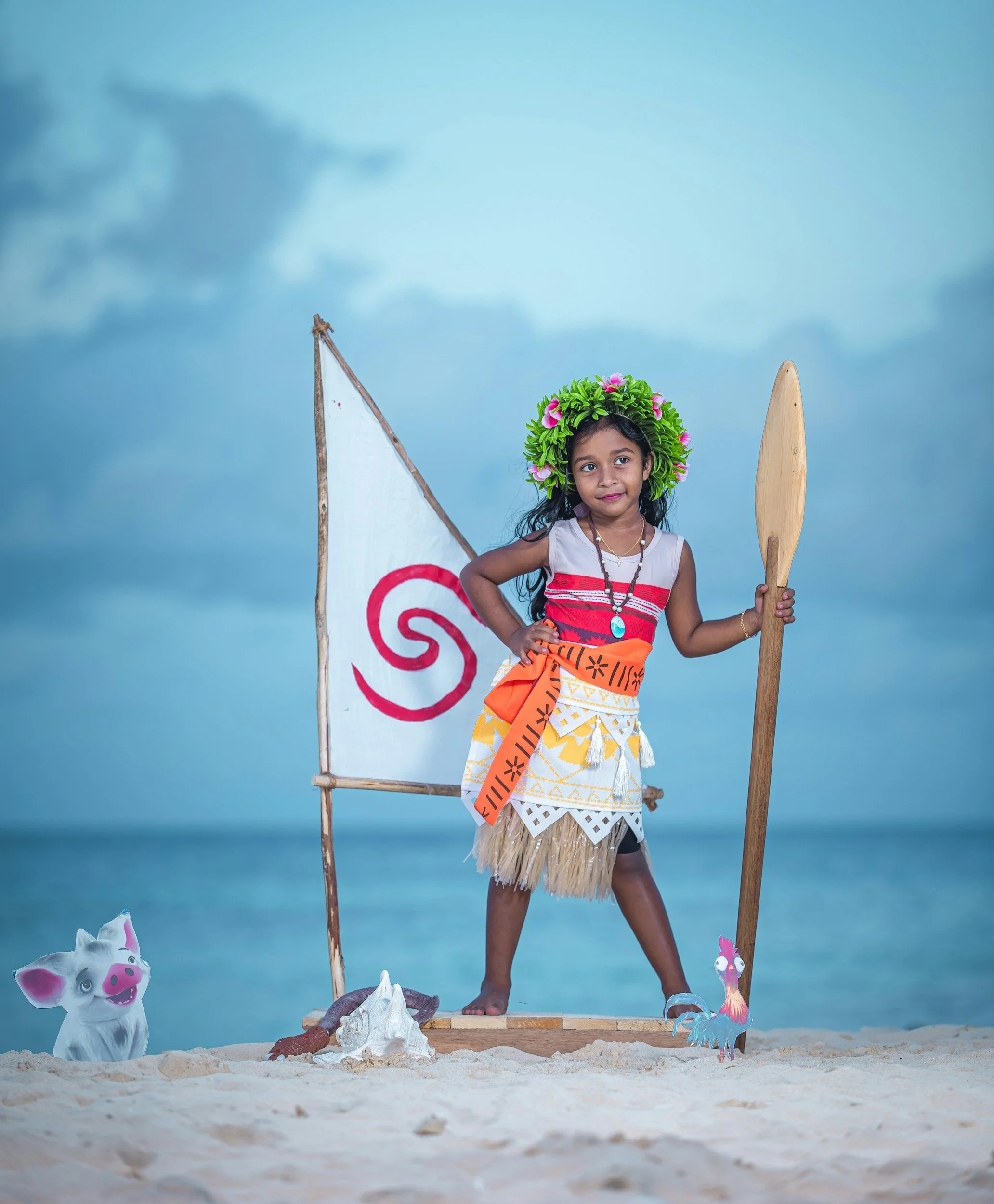 Niña vestida con ropa tradicional caribeña, con corona de flores en la cabeza, sosteniendo una lanza, en una especie de escena teatral en la playa, con fondo de mar y cielo
