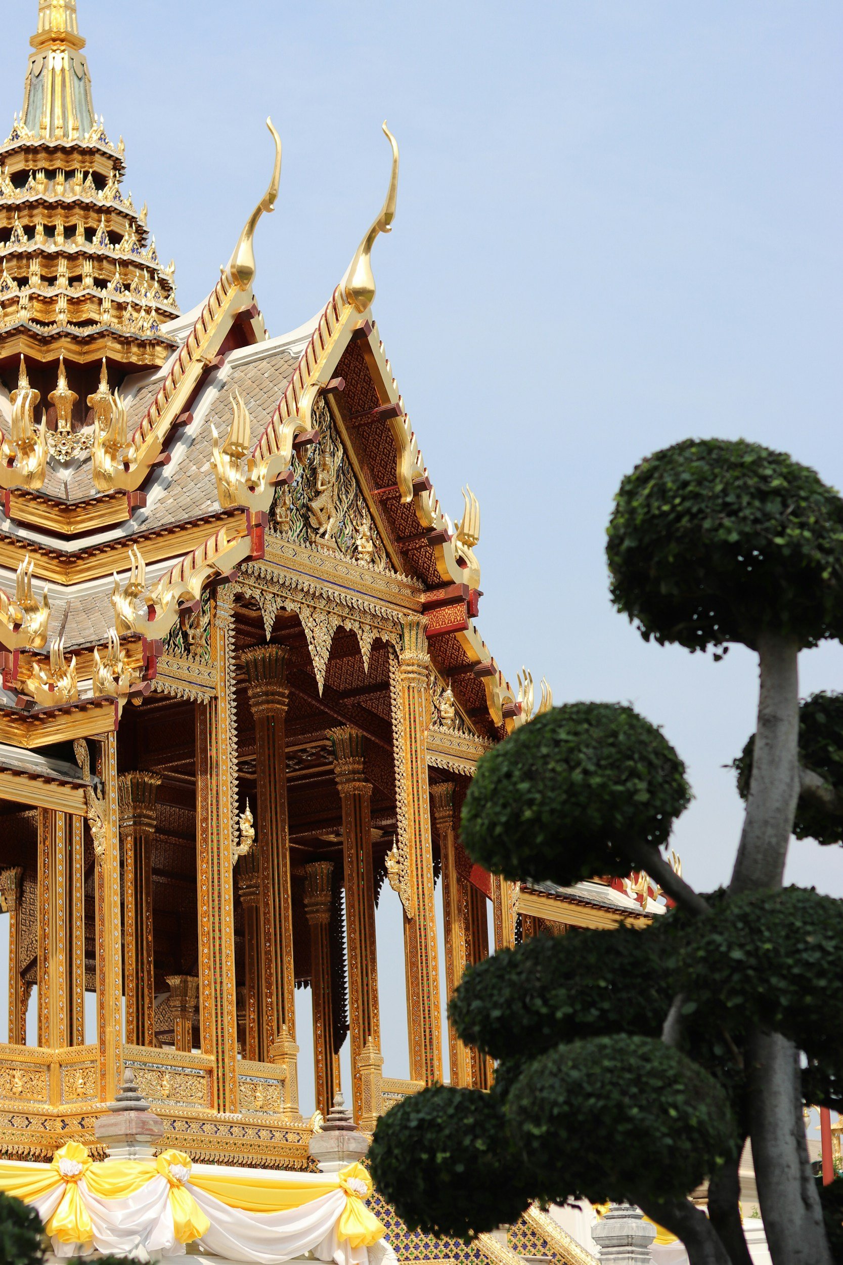 Estructura de un templo tailandés decorado con detalles dorados, rodeada de árboles recortados en forma de nubes y un cielo despejado.
