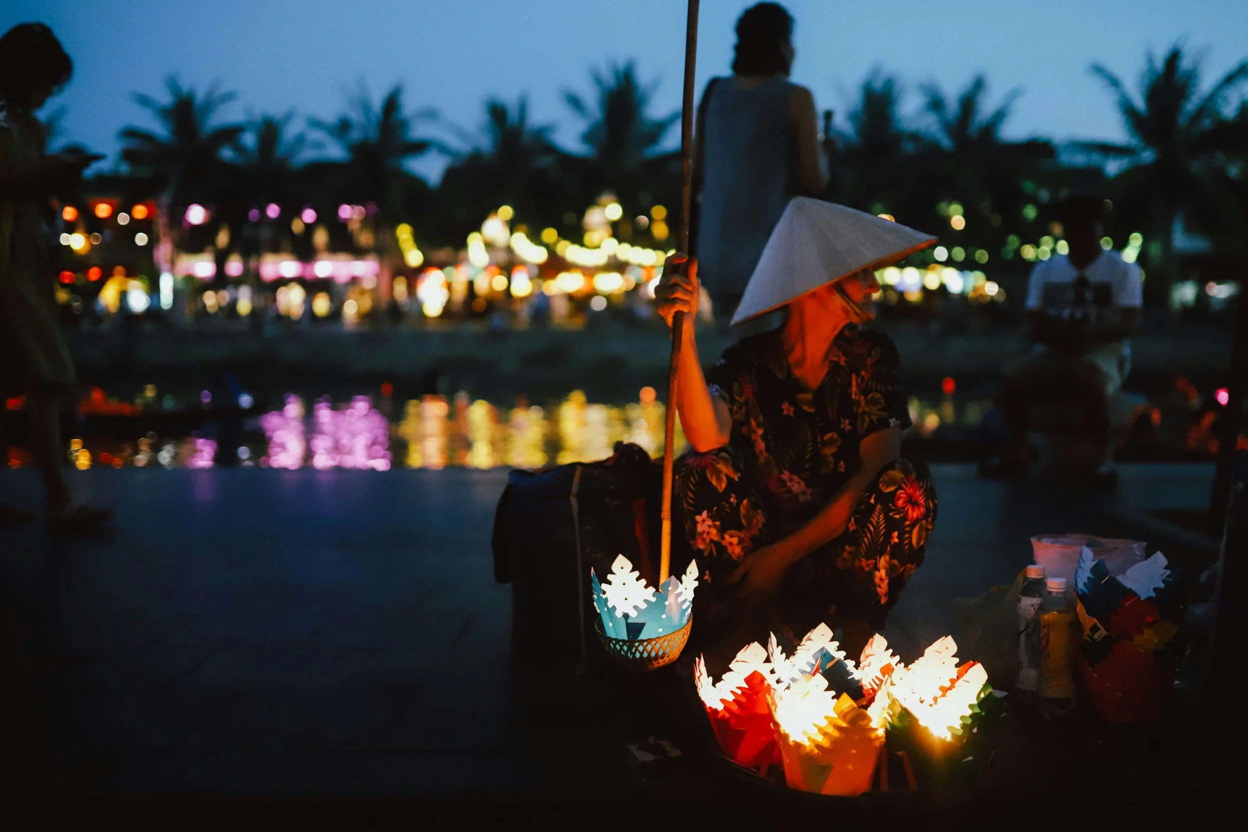 Una persona vendiendo linternas de papel en un mercado nocturno junto al río, en un entorno con palmeras y luces de fondo.