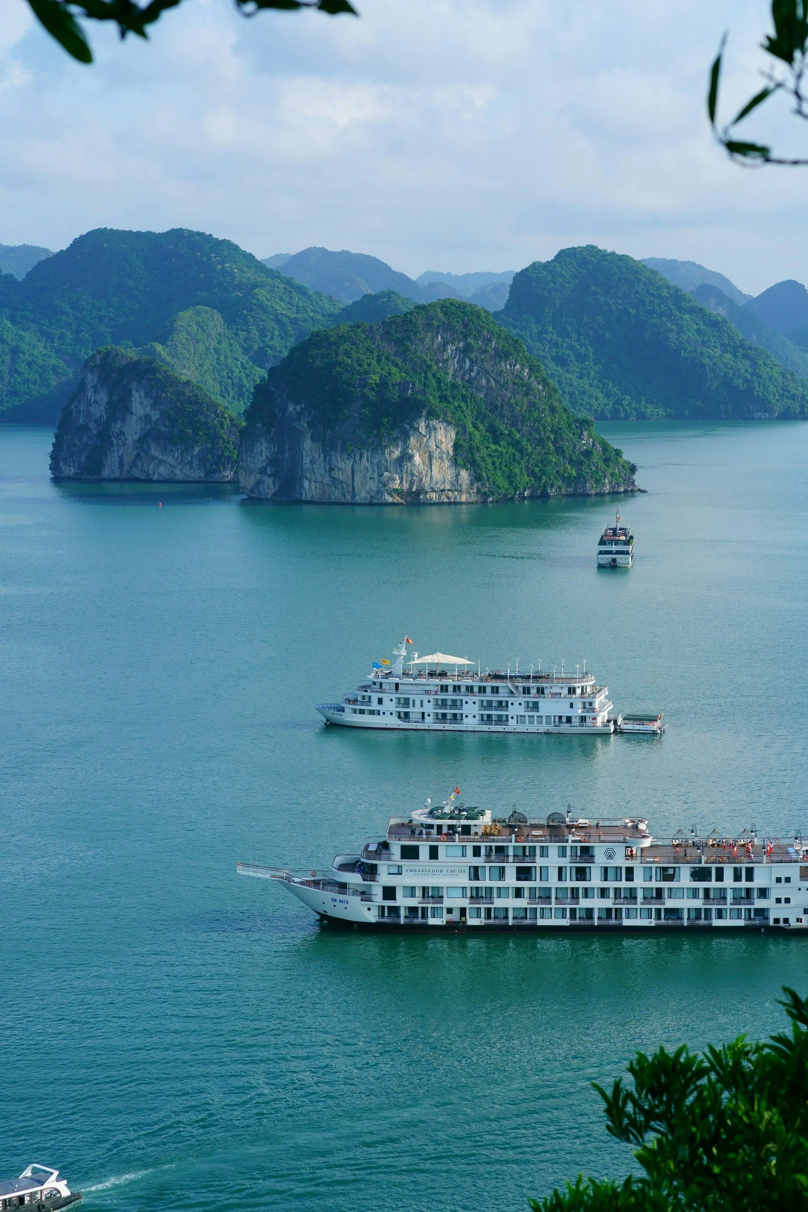 Fotografía de un paisaje marítimo con varias islas rocosas y verdes. Se ven tres barcos grandes navegando en el agua, uno de ellos en primer plano. La escena muestra un ambiente tranquilo con montañas en el fondo y un cielo parcialmente cubierto con nubes.