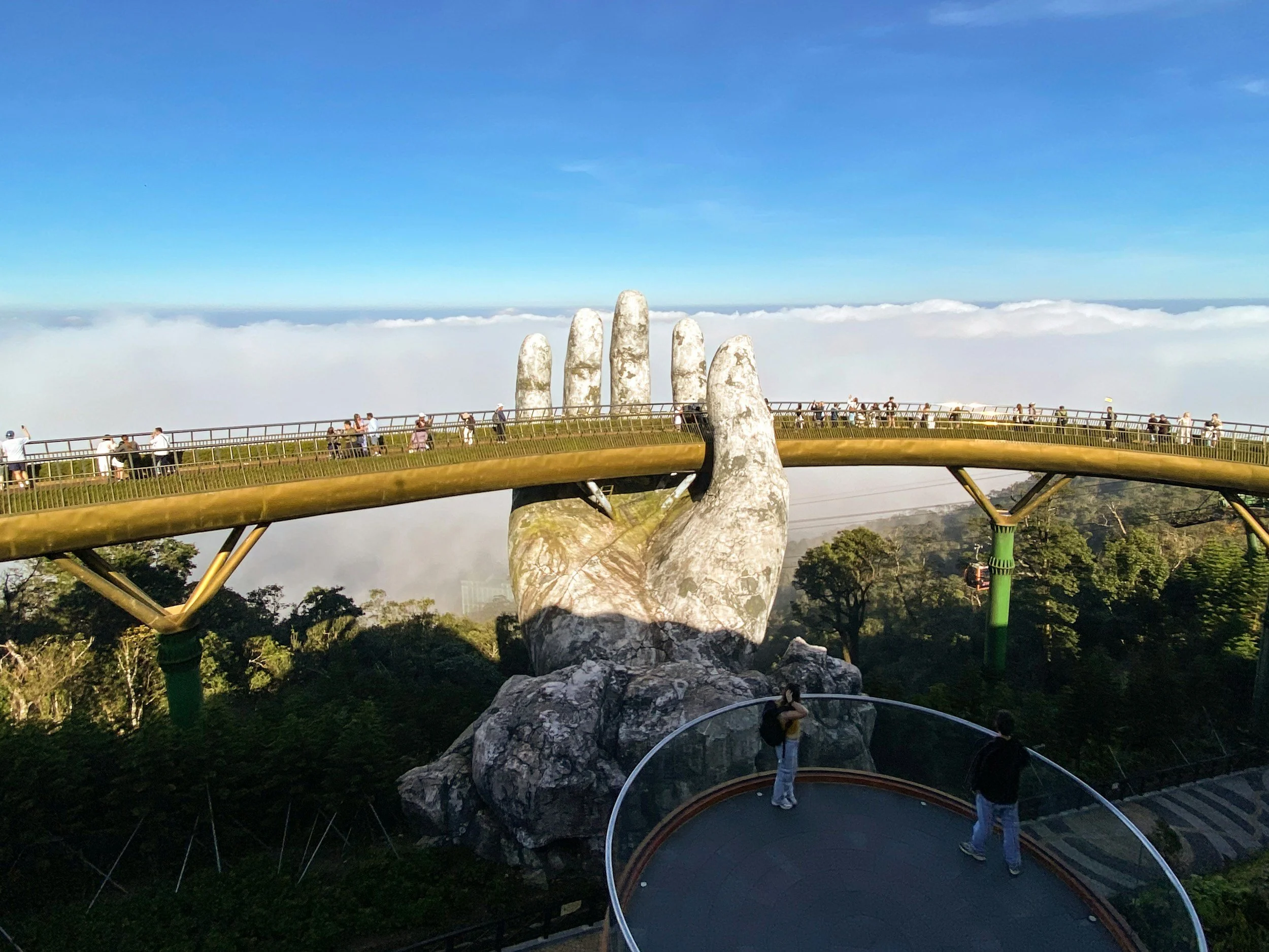 Puente del Corazón en el Monte Huashan en China, con una estructura de mano gigante que sostiene el puente y turistas caminando sobre él, con nubes y cielo azul al fondo.
