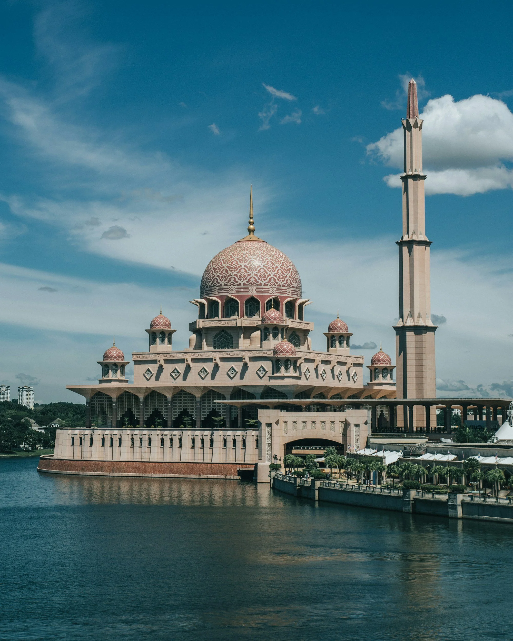 Una mezquita con cúpula grande y varias pequeñas, ubicada junto a un cuerpo de agua, con un cielo azul y algunas nubes.
