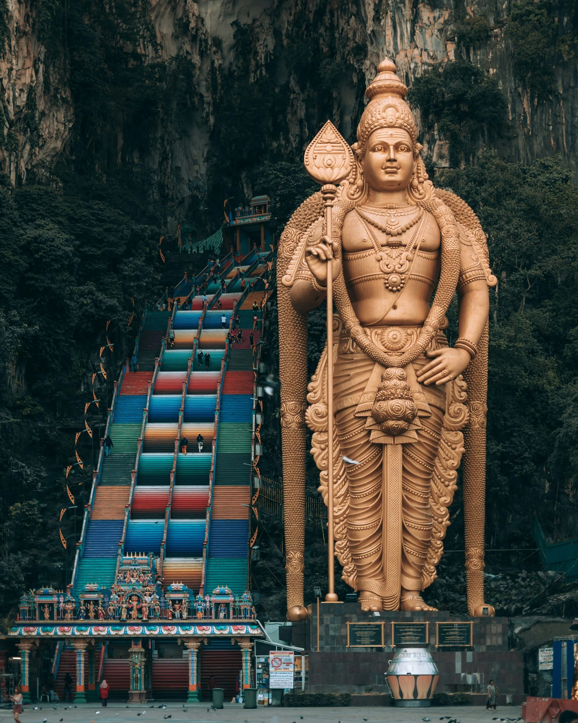 Escalinata de colores en Batu Caves con una gran estatua de Lord Murugan en primer plano, en un entorno natural con vegetación y roca caliza.