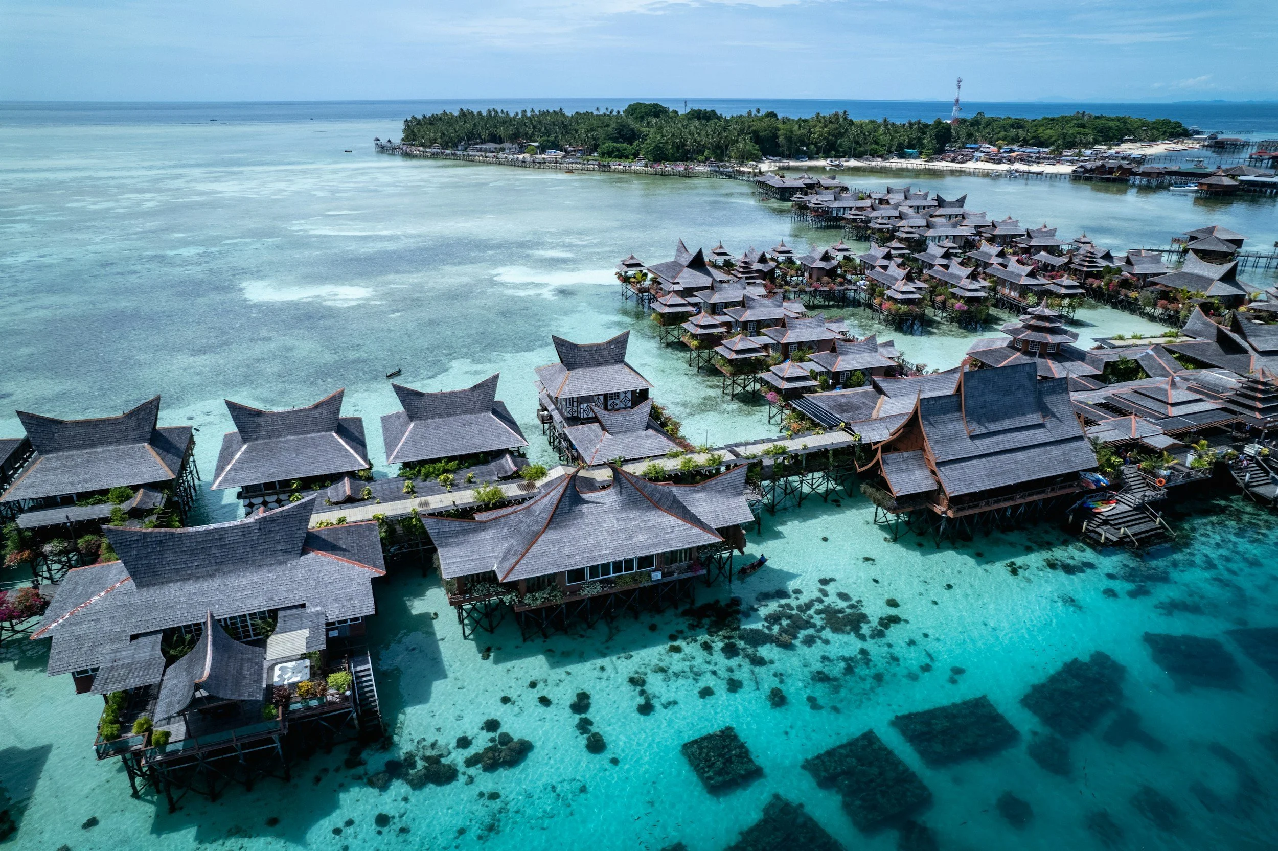 Conjunto de bungalows sobre el agua en una zona costera, con agua cristalina y arrecifes visibles debajo, y una isla con vegetación al fondo.