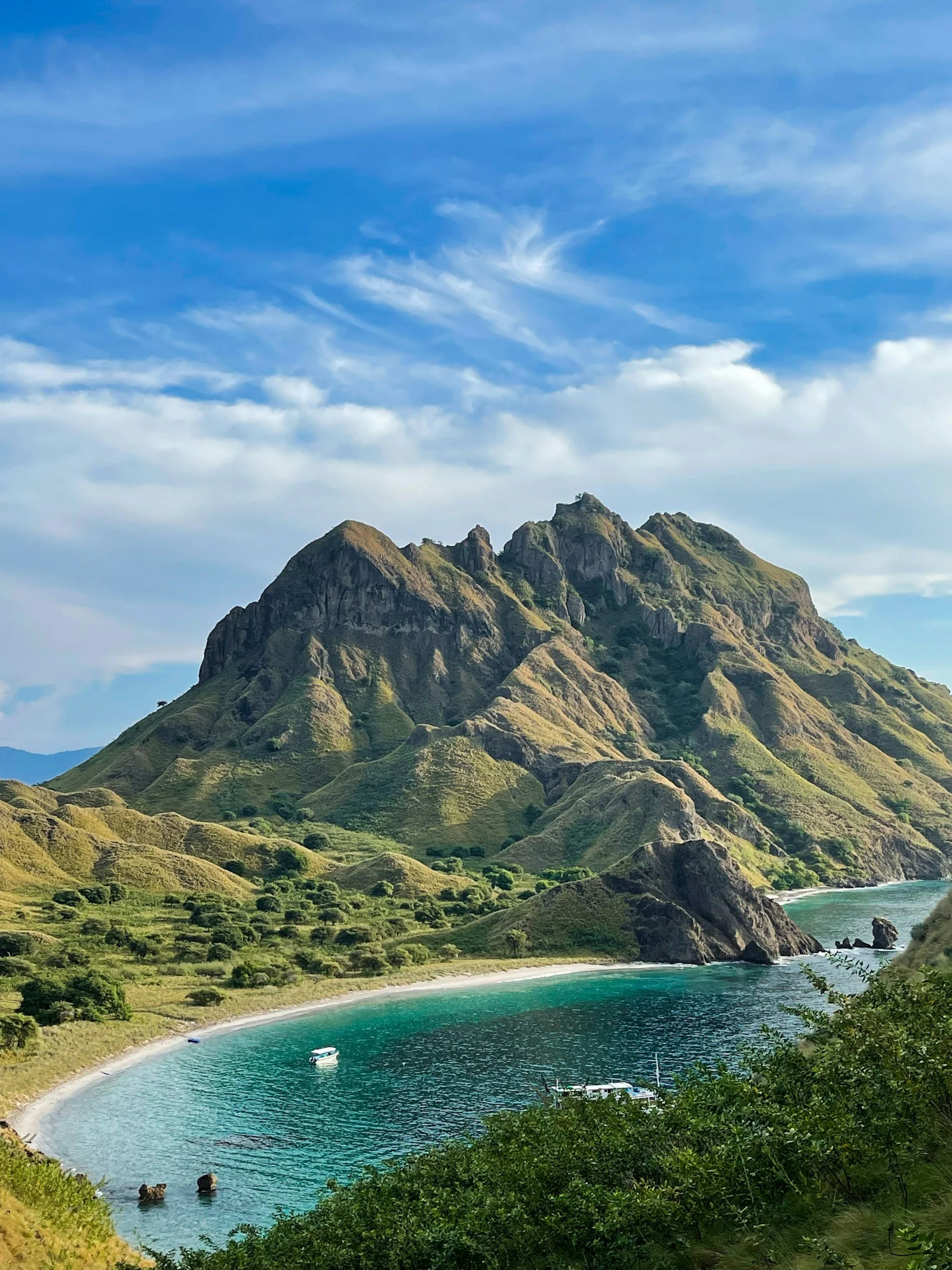 Montaña con pendientes verdes y rocosas, playa de agua azul, cielo con nubes delgadas y barco pequeño en el agua.