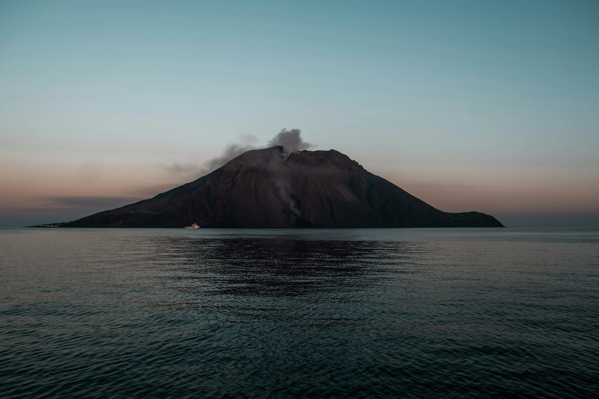 volcán en erupción con humo saliendo de su cráter, frente al mar con reflejo del volcán en el agua