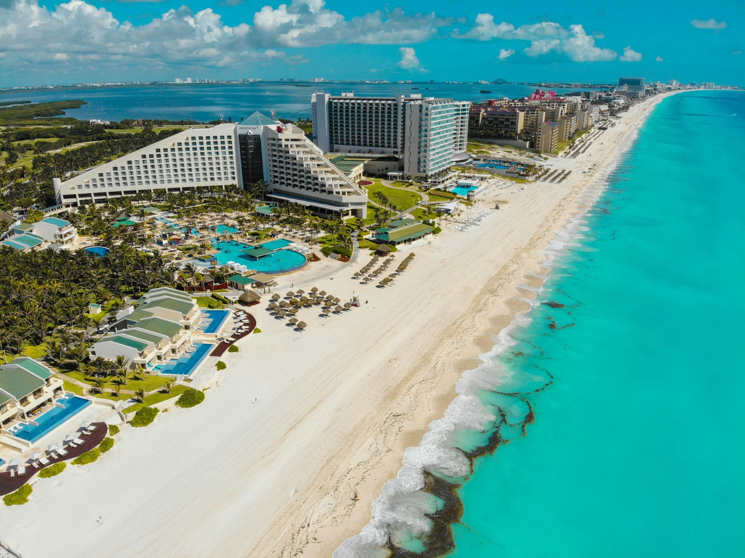 Vista aérea de un hotel en la playa con piscinas, tumbonas y cabanas, junto al mar turquesa, con edificios y cielo con nubes al fondo.