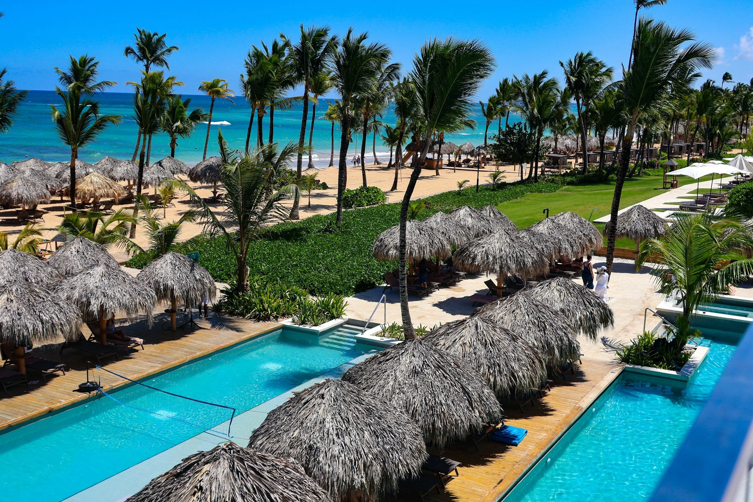 Piscina con sombrillas de paja en primer plano, palmeras y playa con sombrillas y sillas en el fondo, frente al mar azul con cielo despejado.
