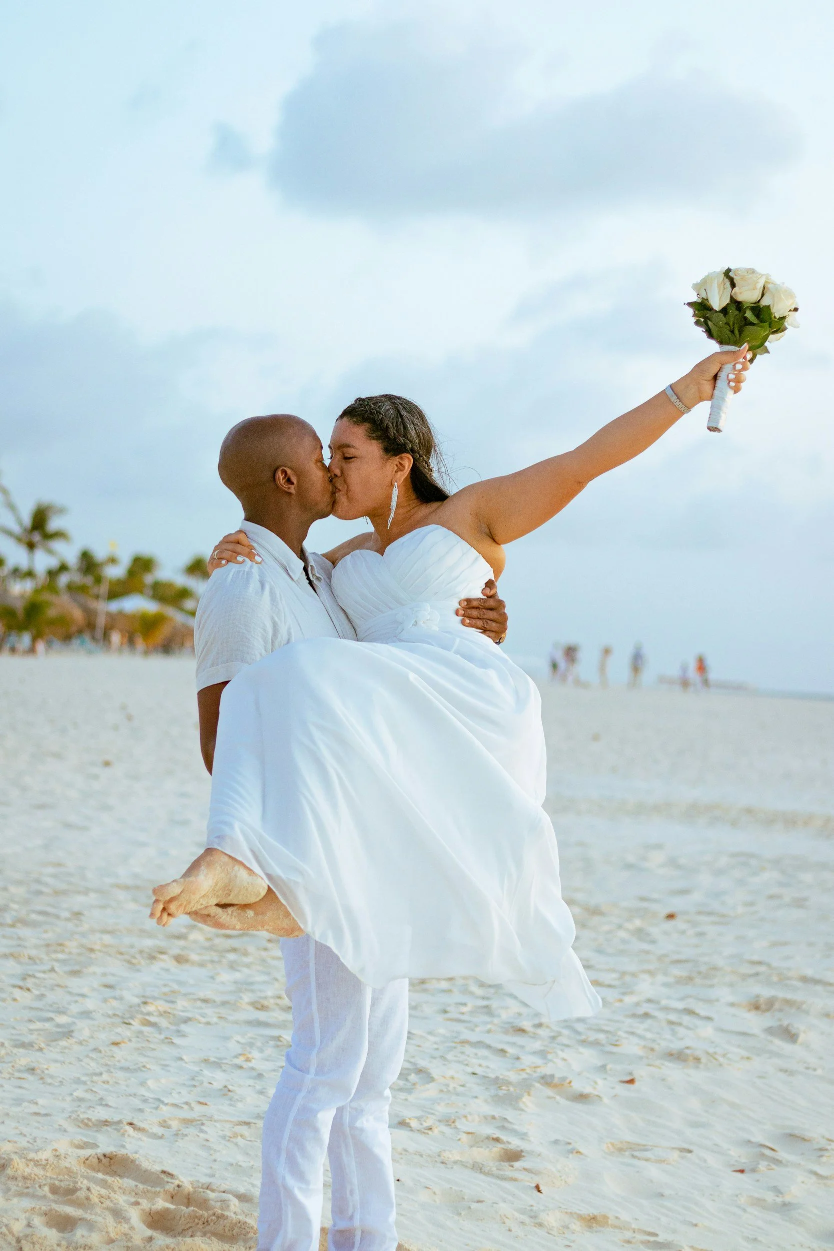 Pareja de novios en la playa, el hombre sostiene a la mujer en sus brazos mientras ambos se besan, ella lleva un vestido blanco y sostiene un ramo de flores blancas en la mano derecha.