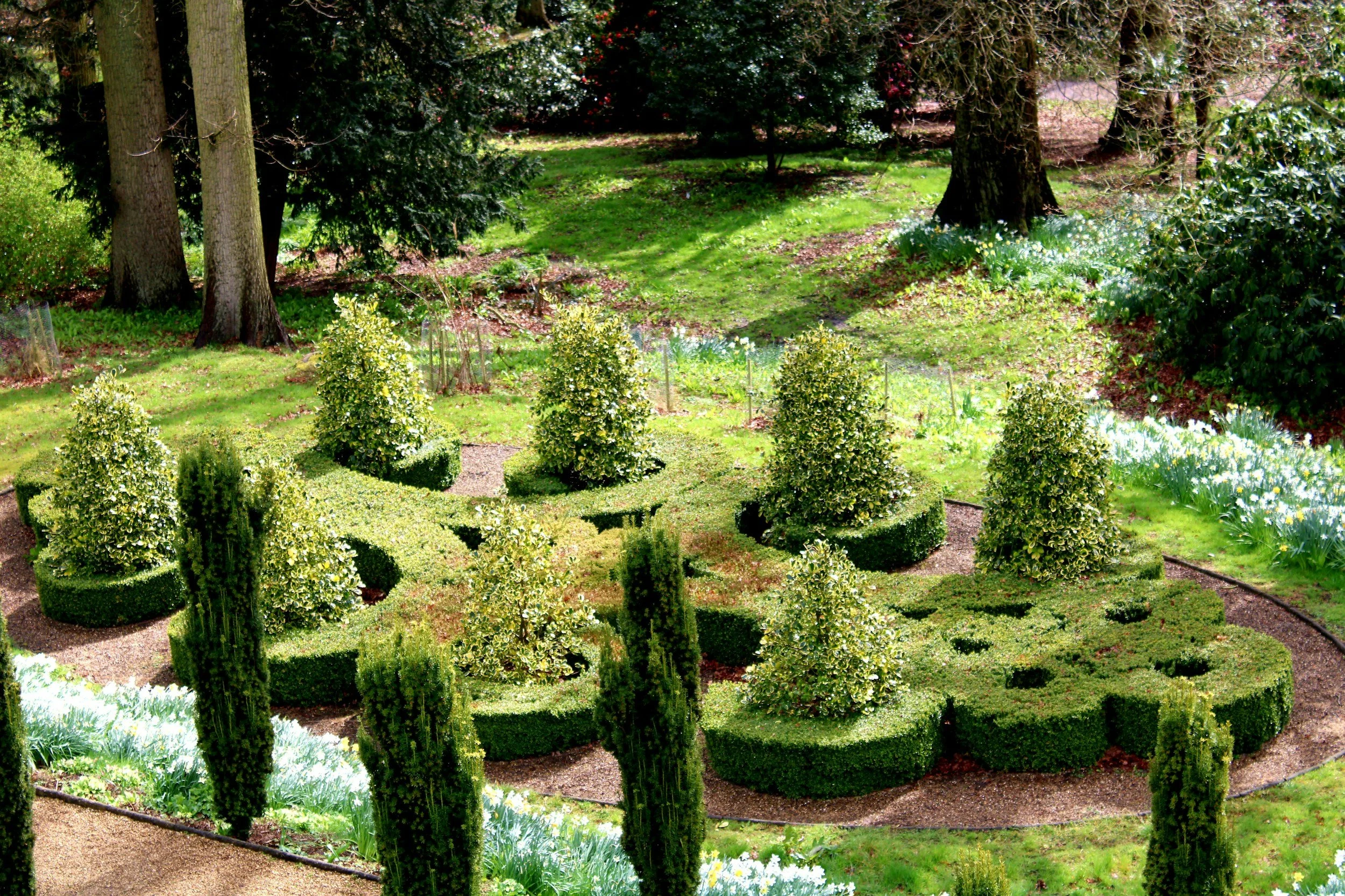 Jardín bien cuidado con arbustos podados en formas geométricas, algunos árboles grandes en el fondo y áreas de césped.