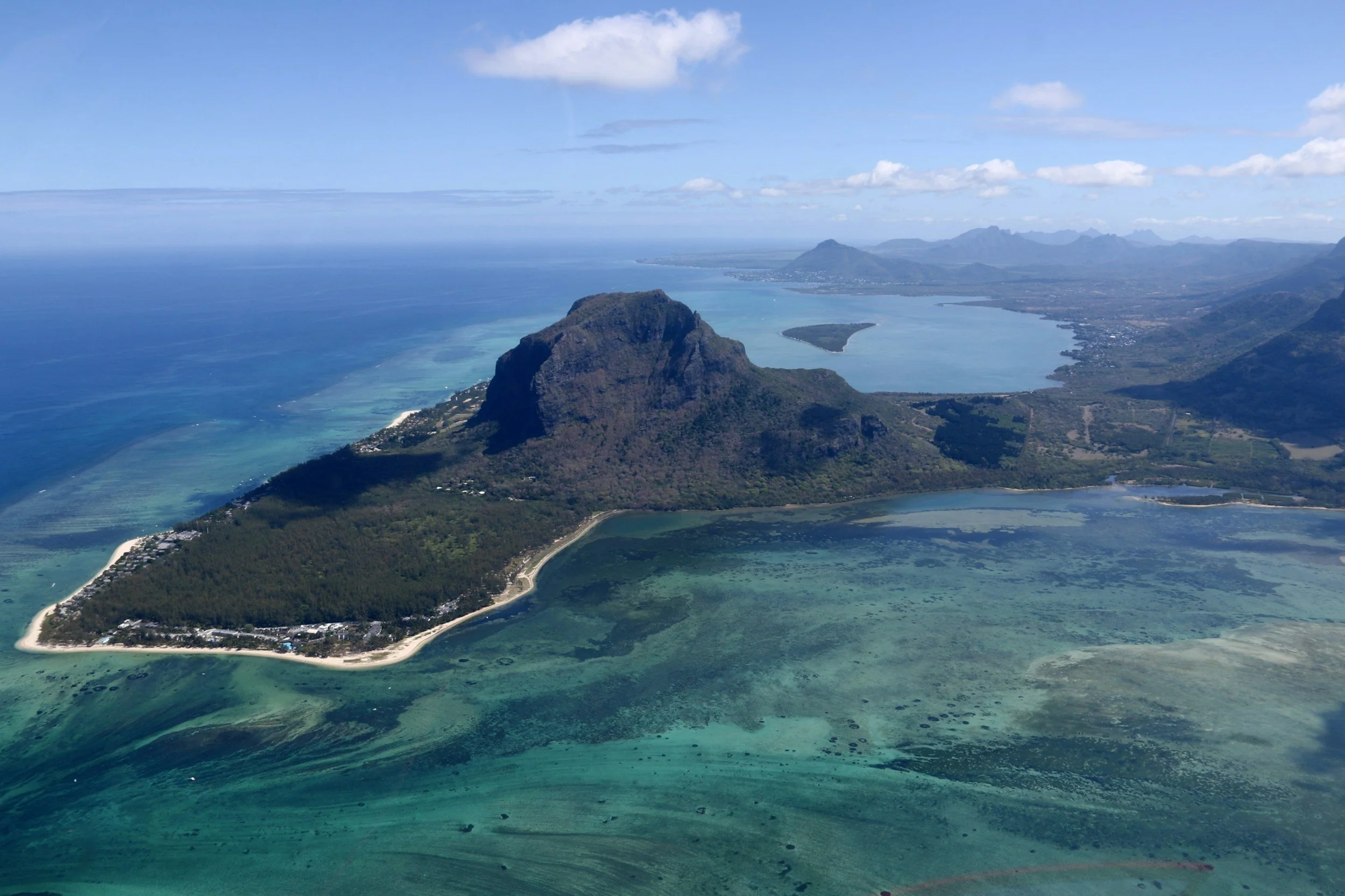 Vista aérea de una isla con montaña central y playas de arena blanca, rodeada de aguas turquesas y un paisaje montañoso en el fondo.