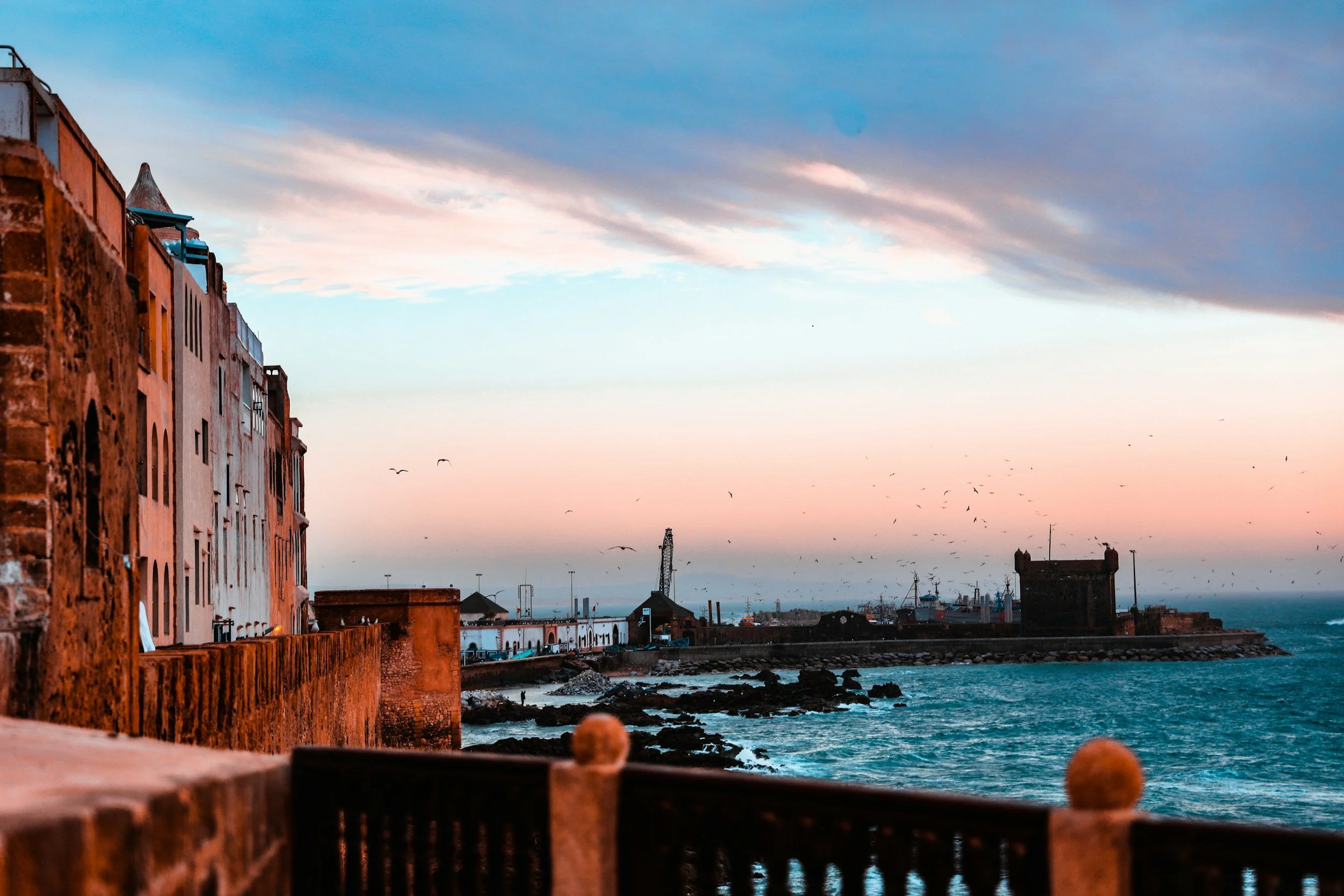 Vista del mar con edificios antiguos a la orilla, cielo con nubes y gaviotas volando, durante el atardecer.