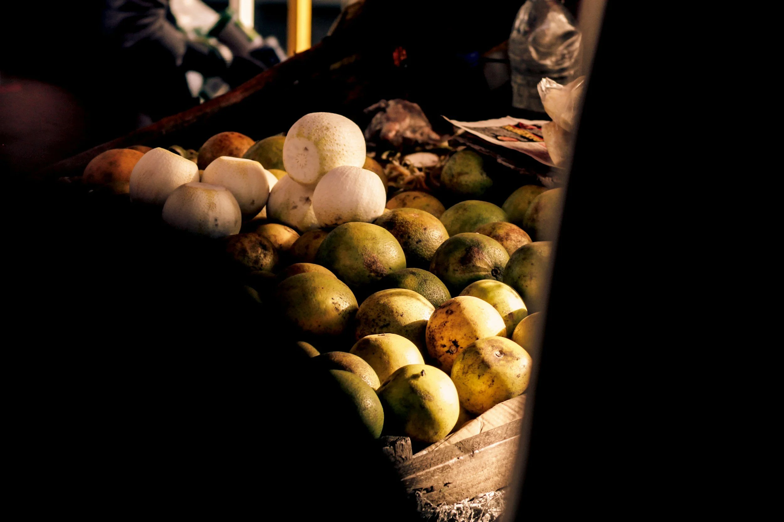 Frutas en un puesto de venta al aire libre, principalmente cocos y guayabas, con luz tenue y sombras.