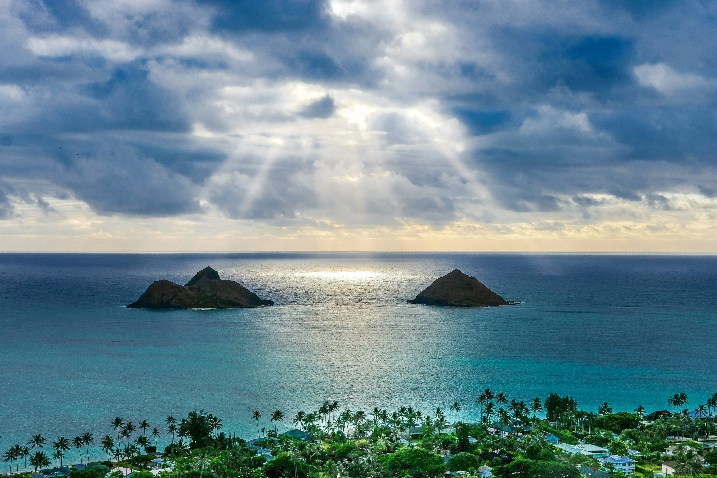 Paisaje costero con dos islas en el océano, nubes en el cielo y rayos de sol atravesando las nubes, vista desde una zona con árboles y casas.