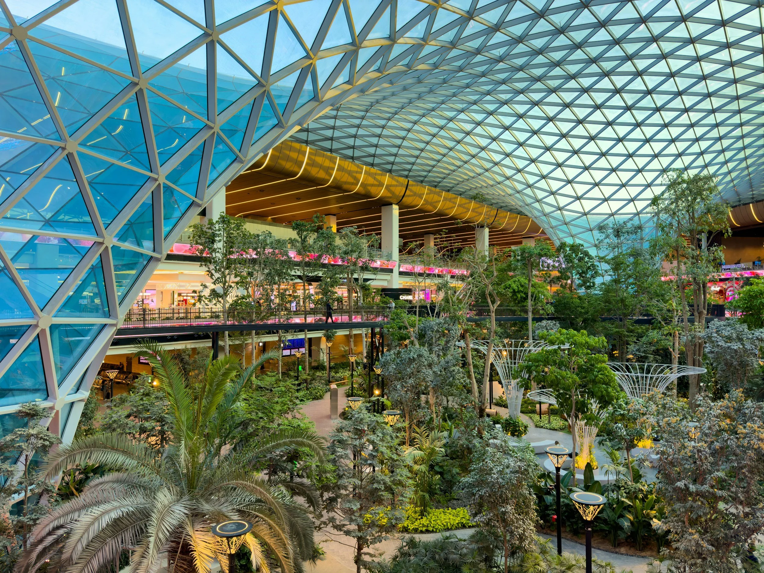 Interior de un centro comercial con un techo de estructura de cristal, árboles y plantas en un espacio abierto, zonas de descanso con farolas y tiendas en diferentes niveles.