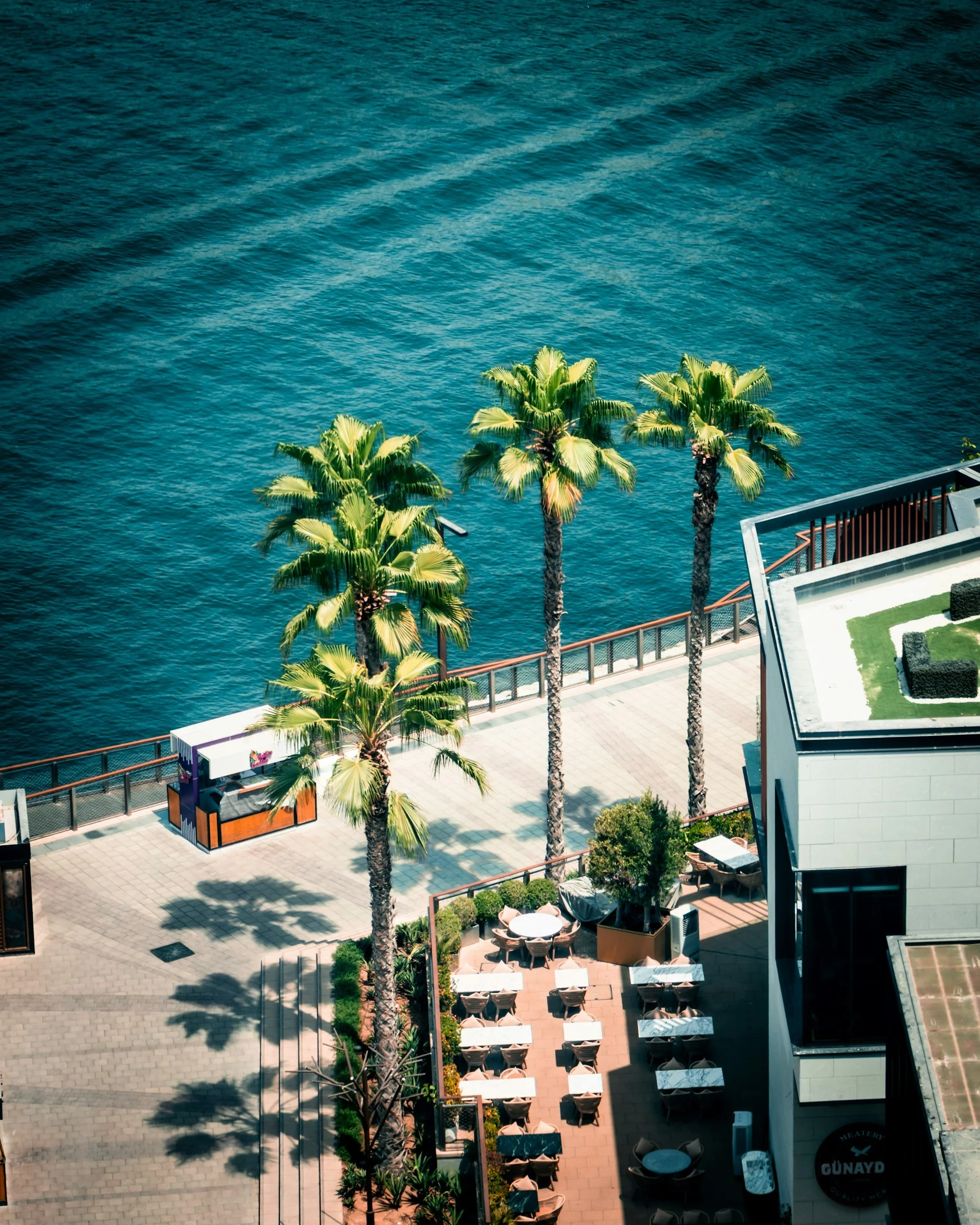Vista aérea de un paseo marítimo con palmeras, mesas de comedor, una estructura de comida rápida y un edificio en la esquina, junto al mar azul.