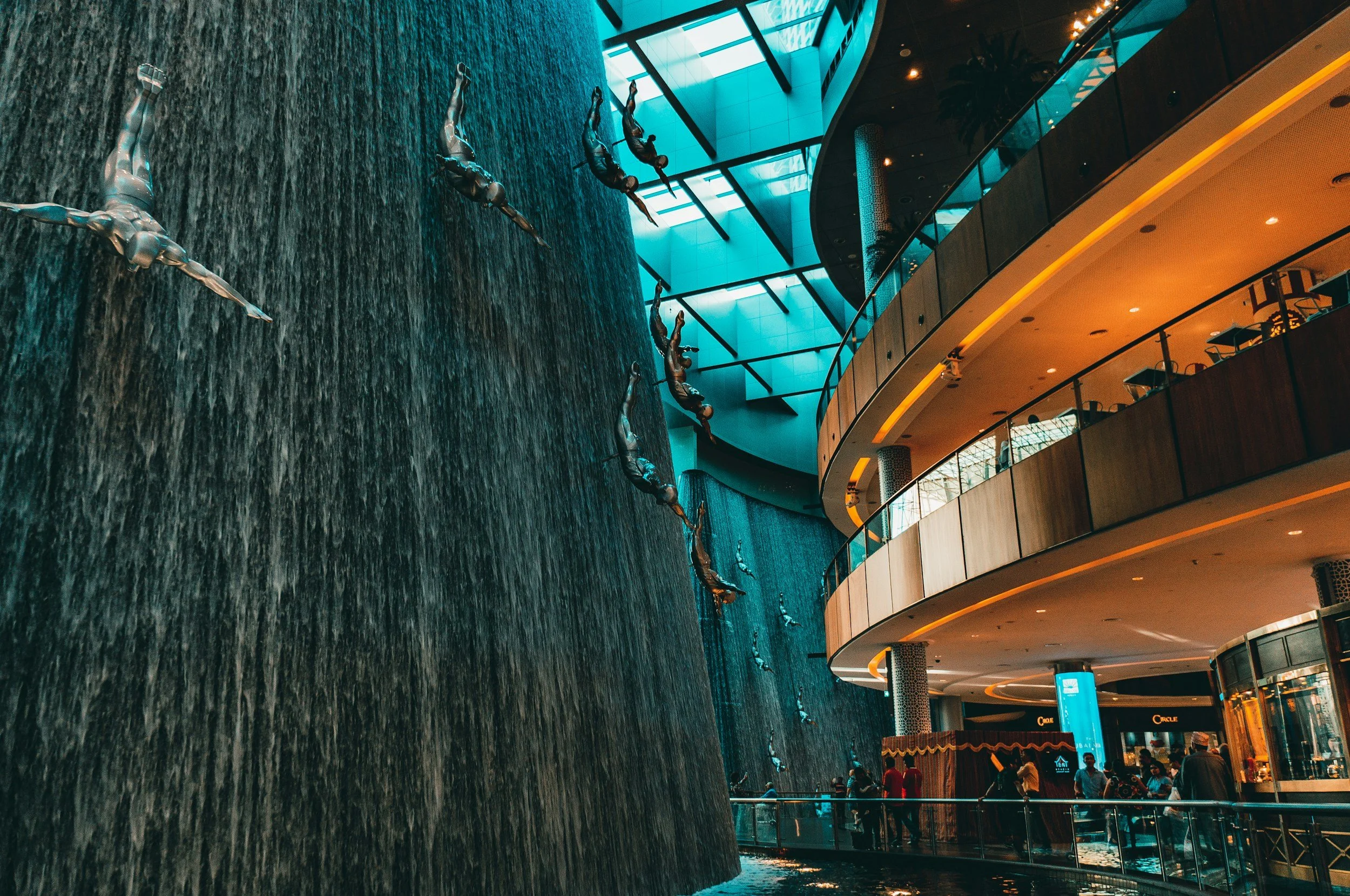 Interiores de un centro comercial con una gran pared de agua decorativa y esculturas de nadadores en la pared, rodeado de varias tiendas y pasillos con personas caminando.