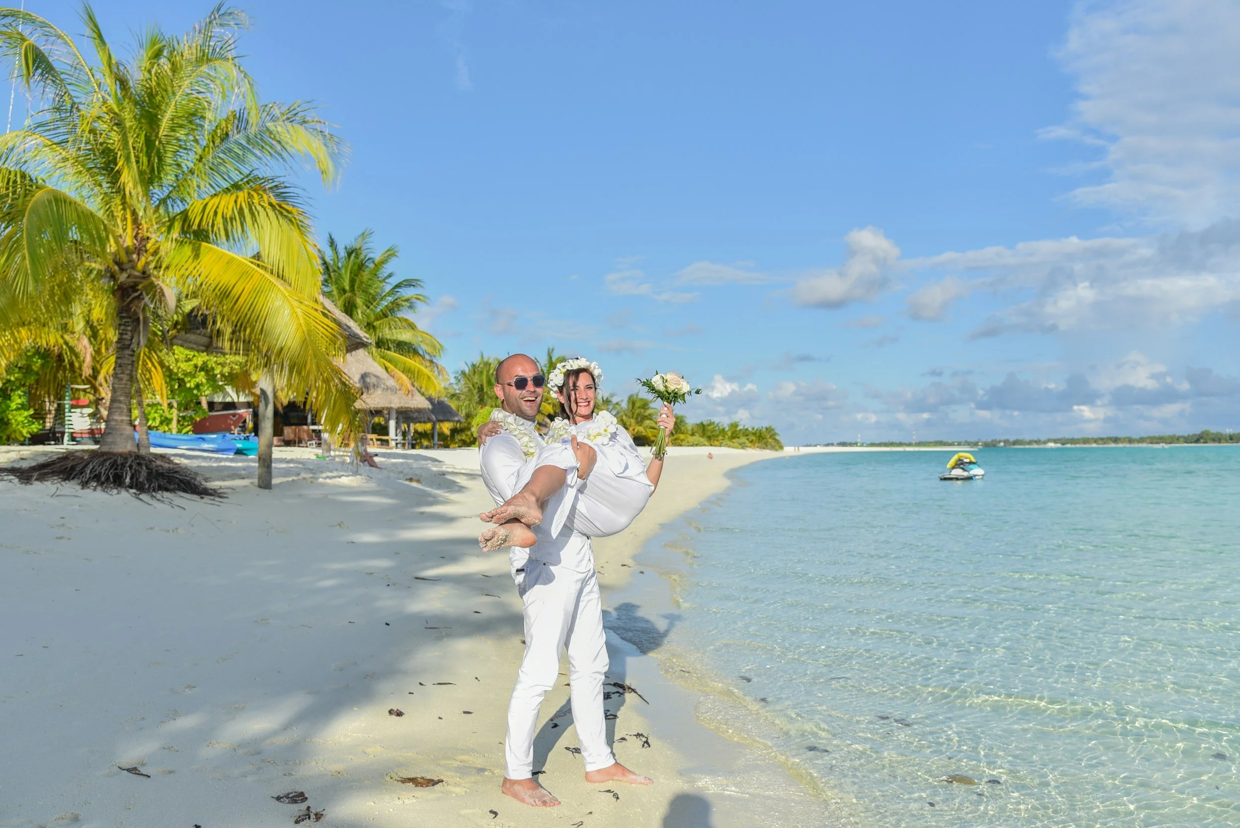 Pareja en ropa de boda en la playa, el hombre carga a la mujer, ambos sonrientes, con flores y fondo de palmeras y mar.