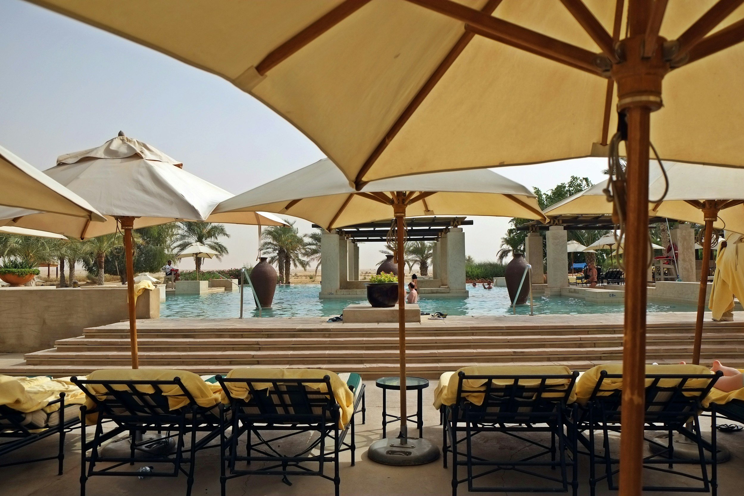 Poolside scene with lounge chairs and large umbrellas, pool with people, palm trees, and decorative vases in a sunny outdoor setting.