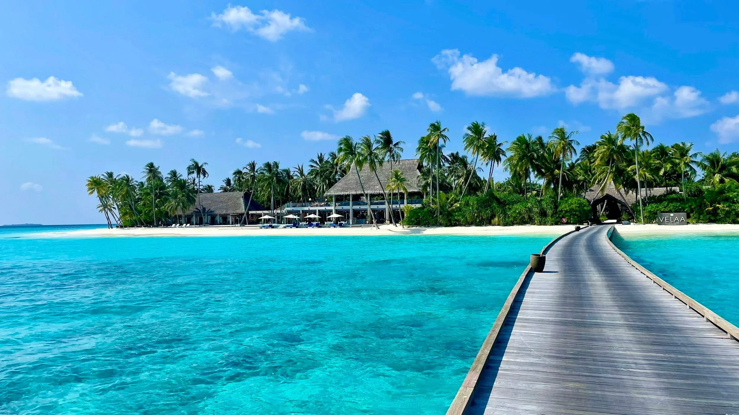 A tropical island resort with a wooden pier leading to a building surrounded by palm trees, turquoise water, and a blue sky with some clouds.