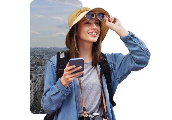 Joven mujer sonriendo, usando sombrero y gafas, con mochila y cargando un teléfono, en un entorno urbano.