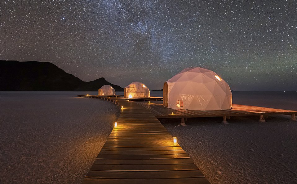 Tres cúpulas geodésicas iluminadas en una playa bajo un cielo estrellado, con una pasarela de madera que conecta las cúpulas y varias velas encendidas a lo largo del camino.
