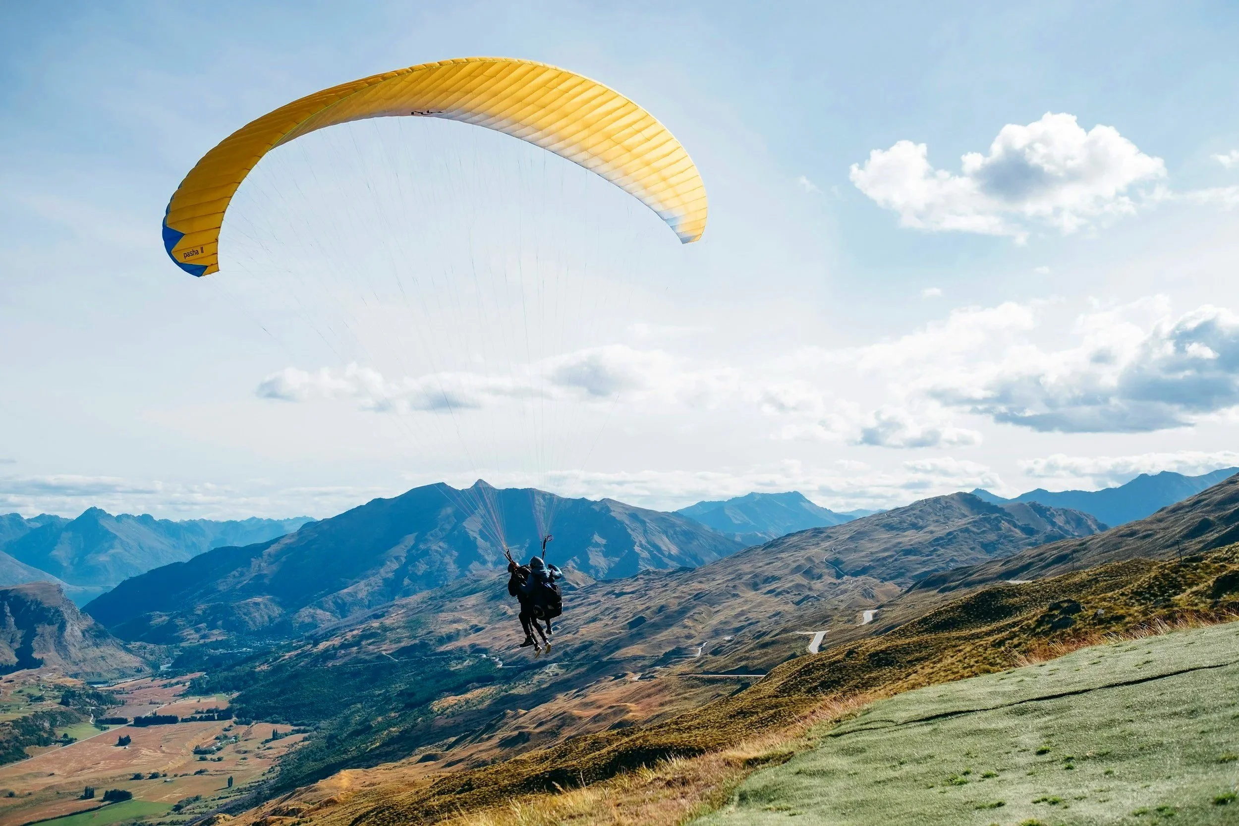 Persona en parapente sobre un paisaje montañoso con cielo despejado y nubes blancas.