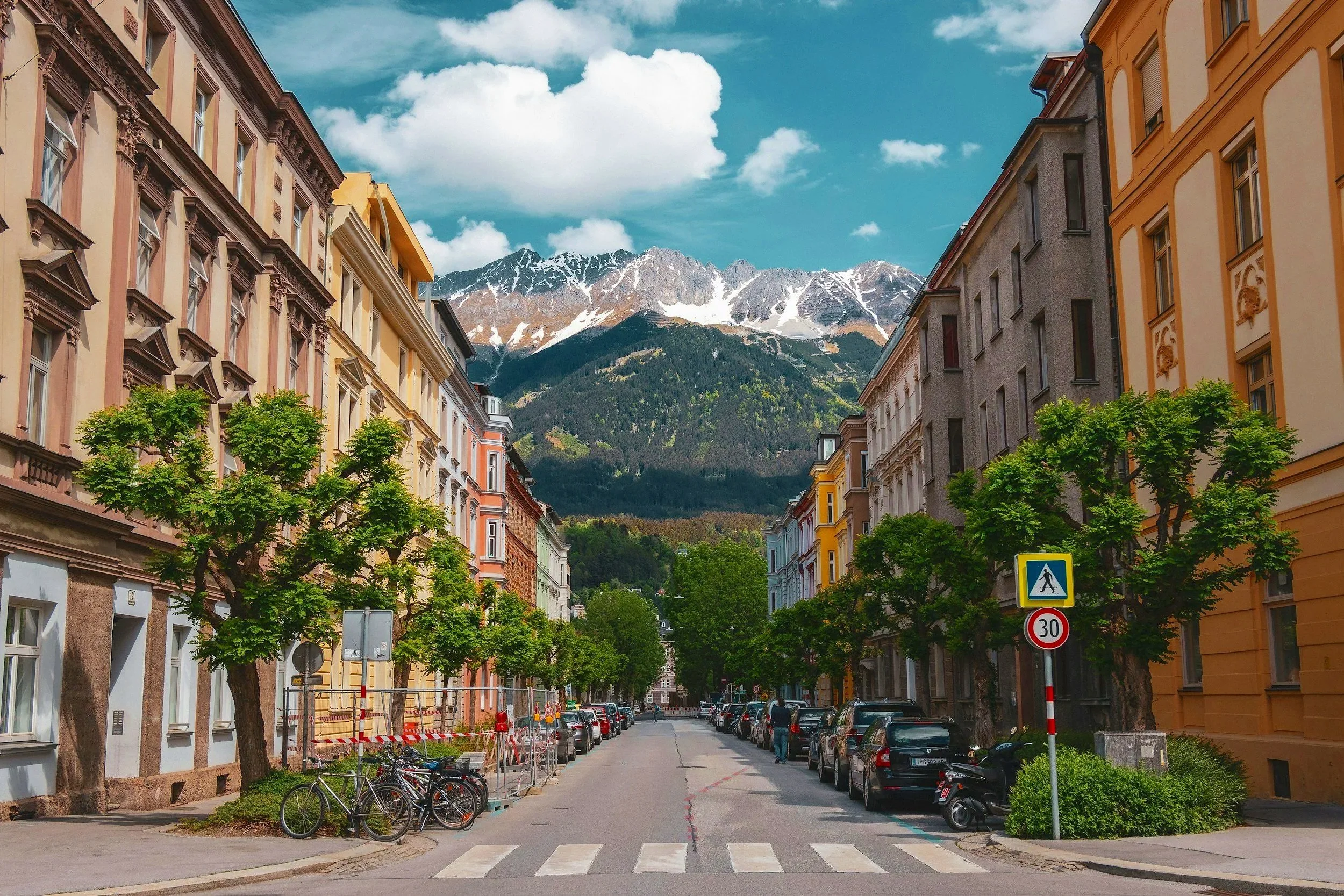 Vista de una calle en una ciudad con edificios coloridos, árboles a los lados, autos estacionados y montañas nevadas al fondo bajo un cielo azul con algunas nubes.