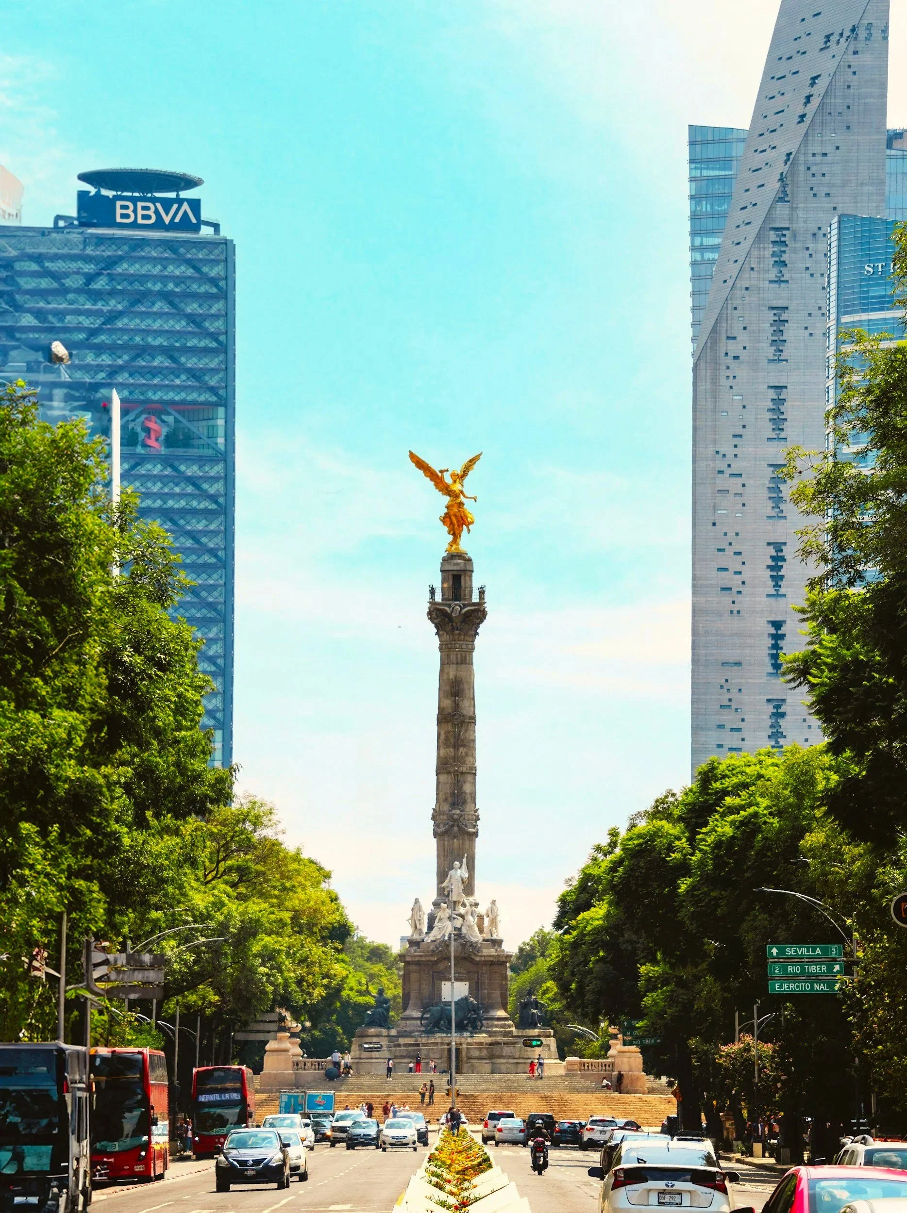 Monumento a la independencia en la Ciudad de México con edificios modernos de fondo y árboles a los lados, tráfico de autos y autobuses en primer plano.